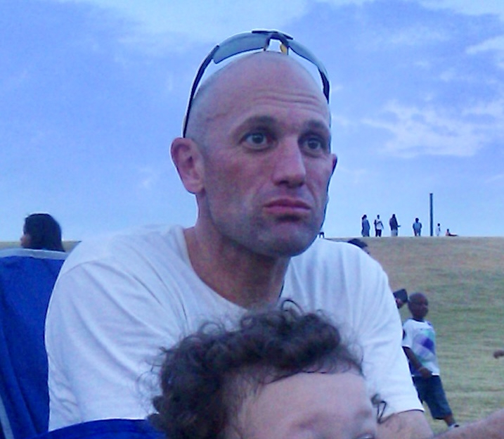 Bald man with sunglasses on head, wearing a white shirt, outdoors with a grassy hill and people in background.