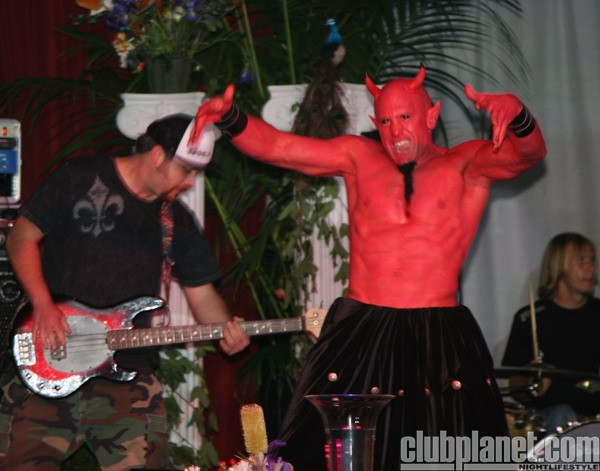 Person dressed as a devil with red body paint, horns, and black skirt, flexing muscles on stage with a guitarist nearby.