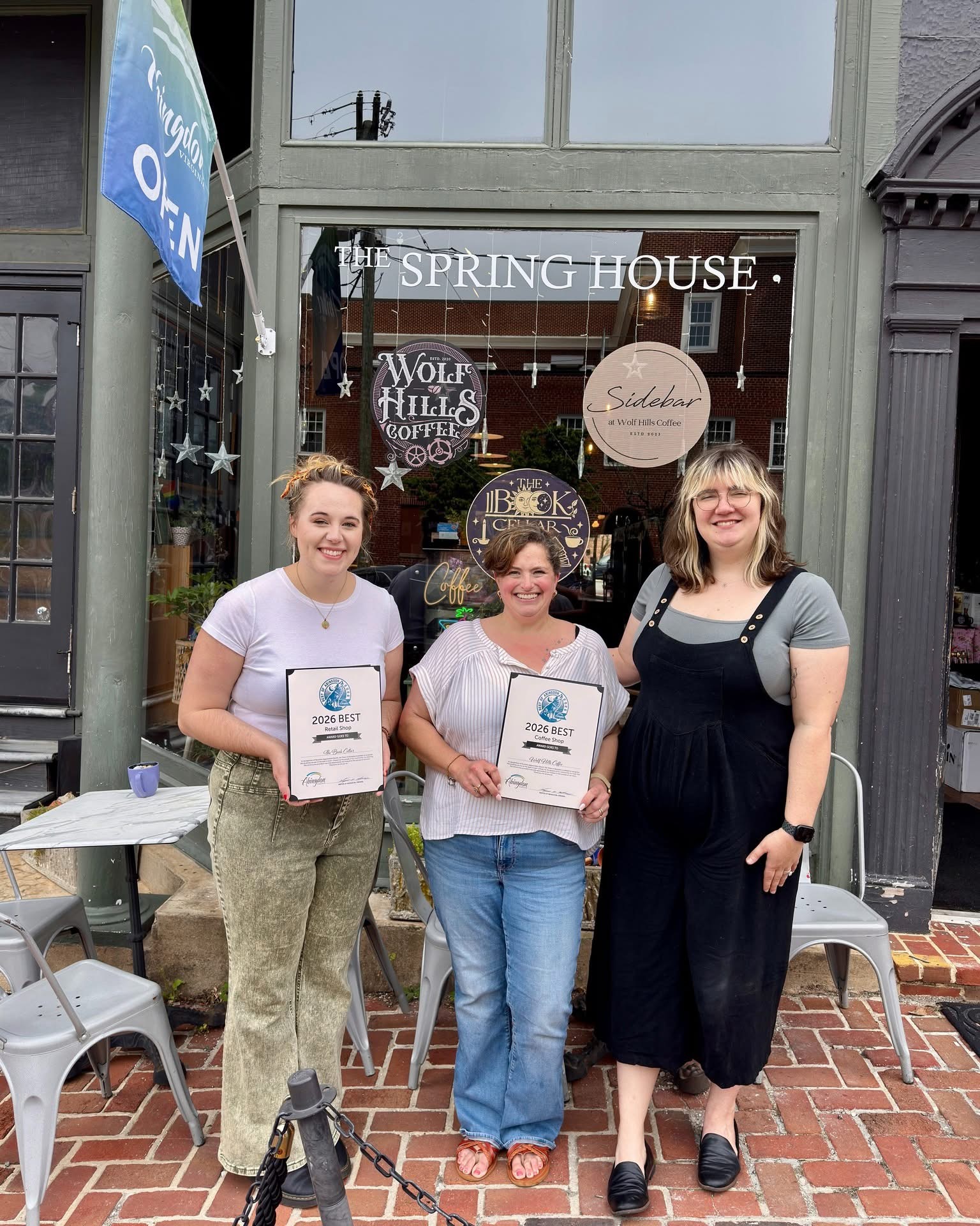 Three women standing outside a shop, holding certificates, smiling, with tables and chairs nearby.