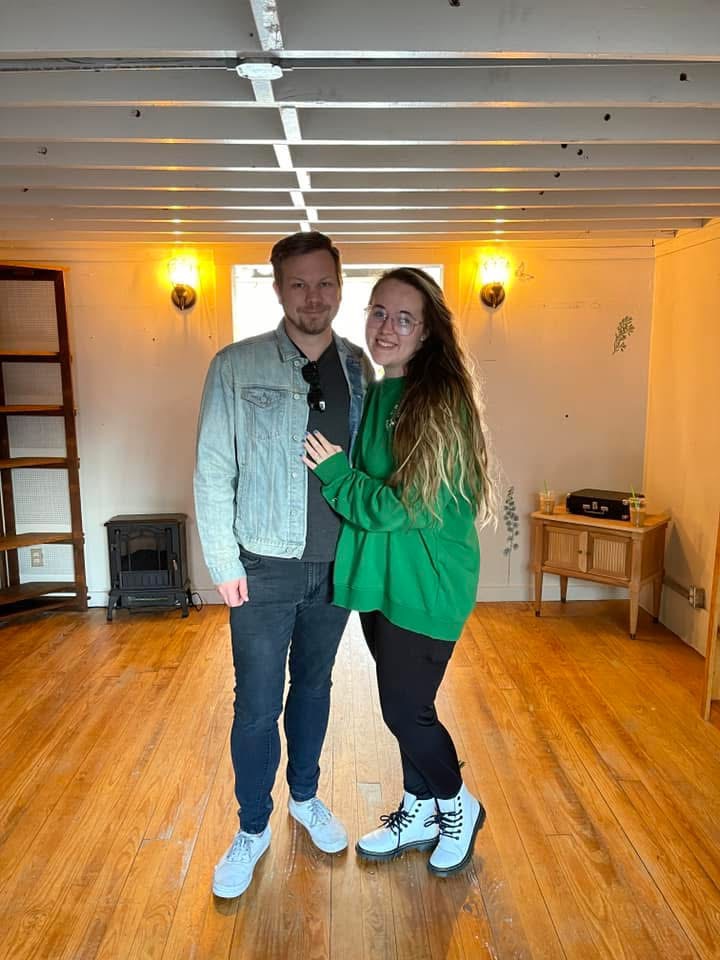 Two people standing indoors, smiling, with wooden floor and wall lights behind them.