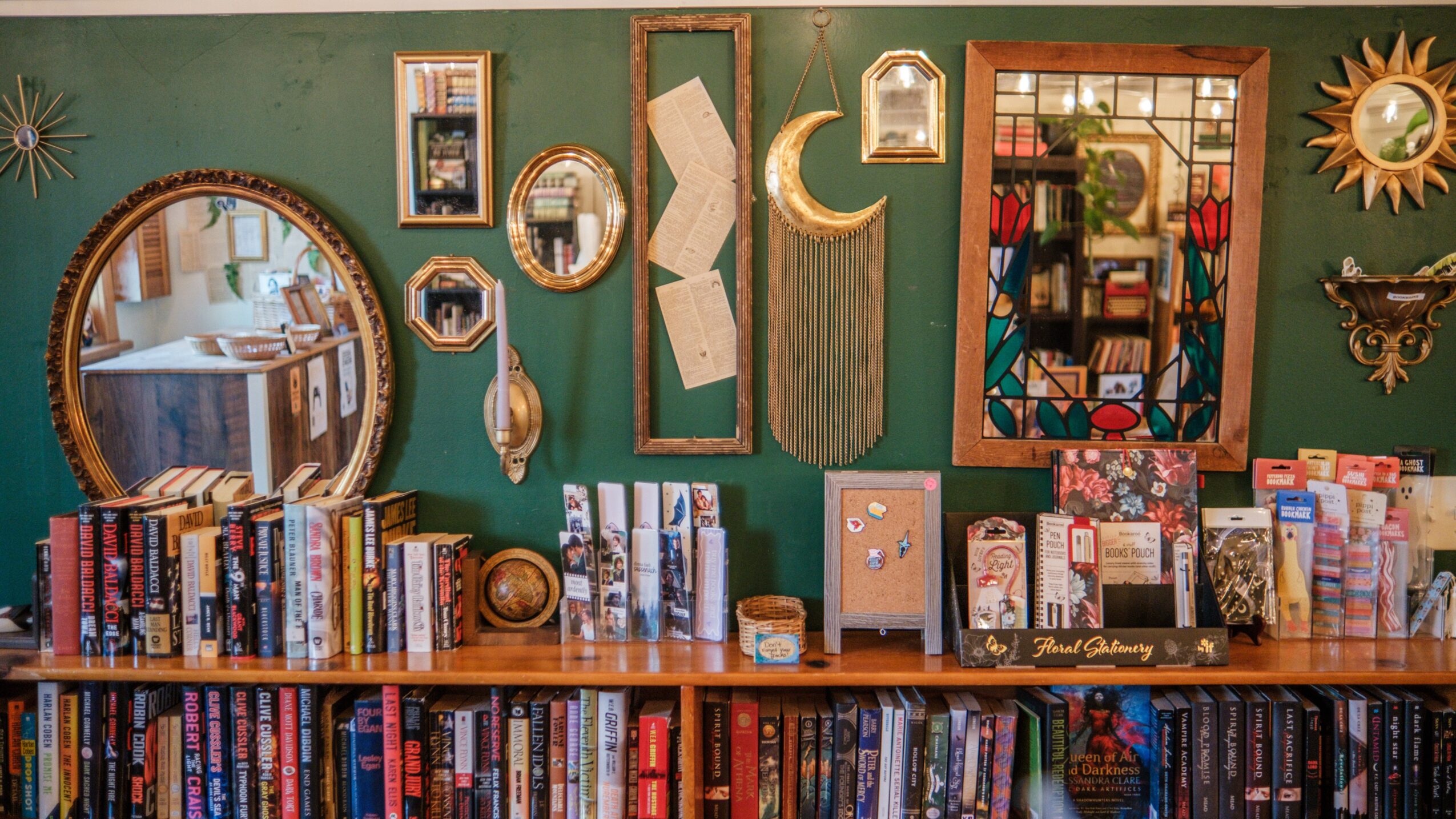 Bookshelves with framed mirrors and decorative items against a green wall, with a wooden table below filled with books.