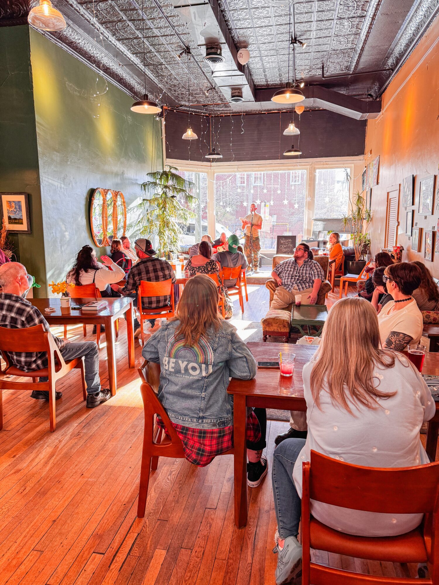People seated at tables in a brightly lit cafe with large windows, colorful walls, and a person speaking at the front.