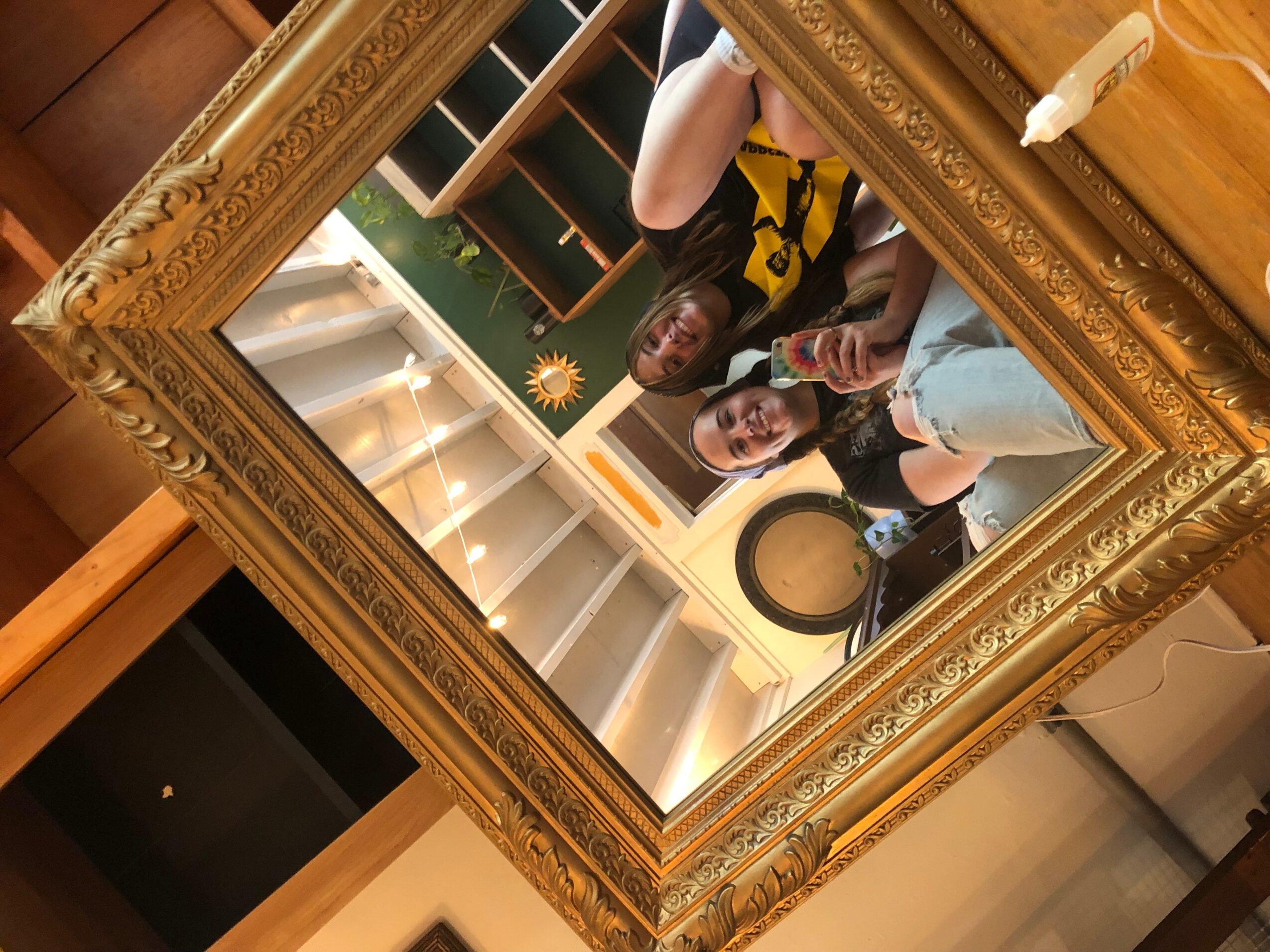 Two people taking a selfie in a large ornate mirror, with a bookshelf and a sun decoration in the background.