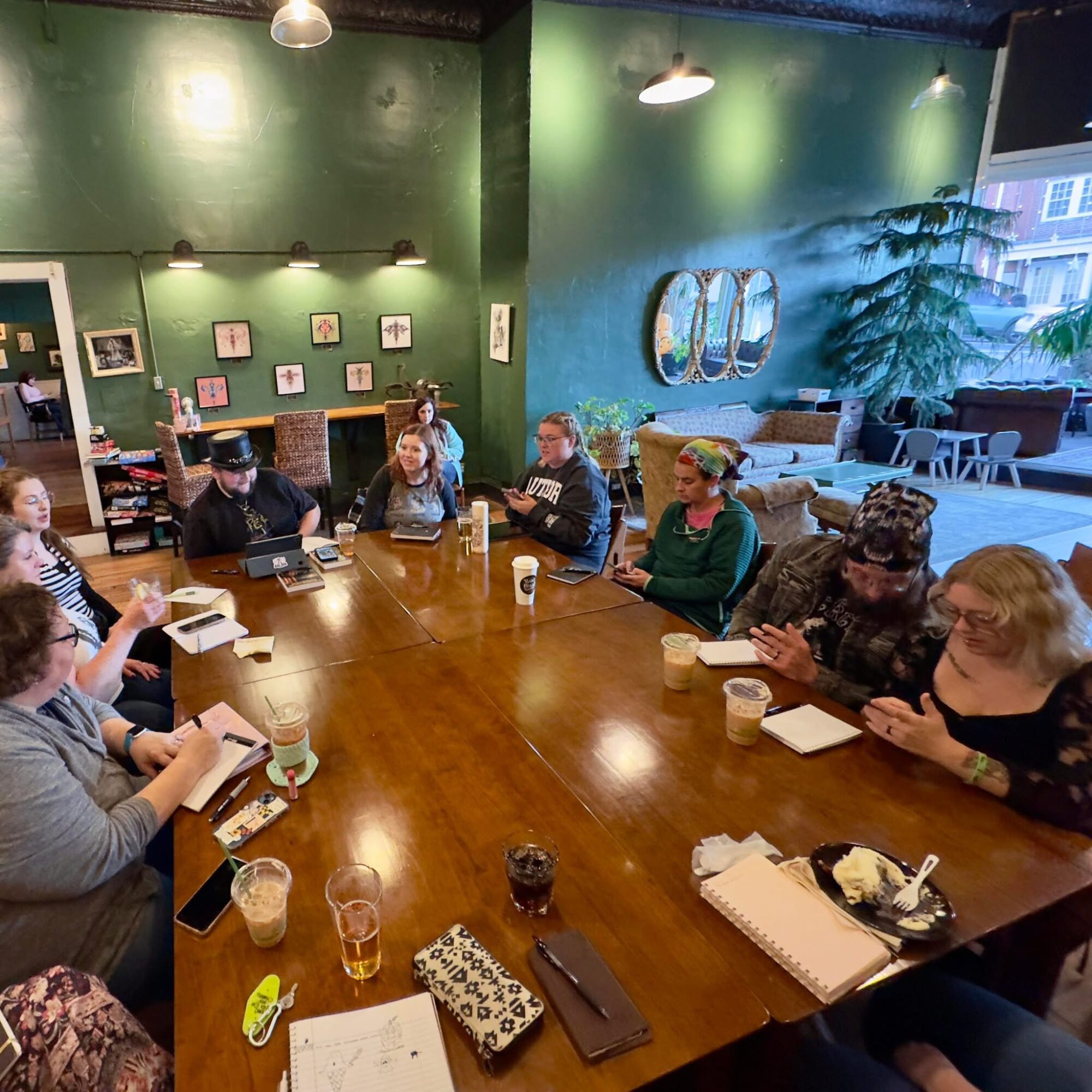 People sitting around a large wooden table in a cozy cafe with green walls and large windows.