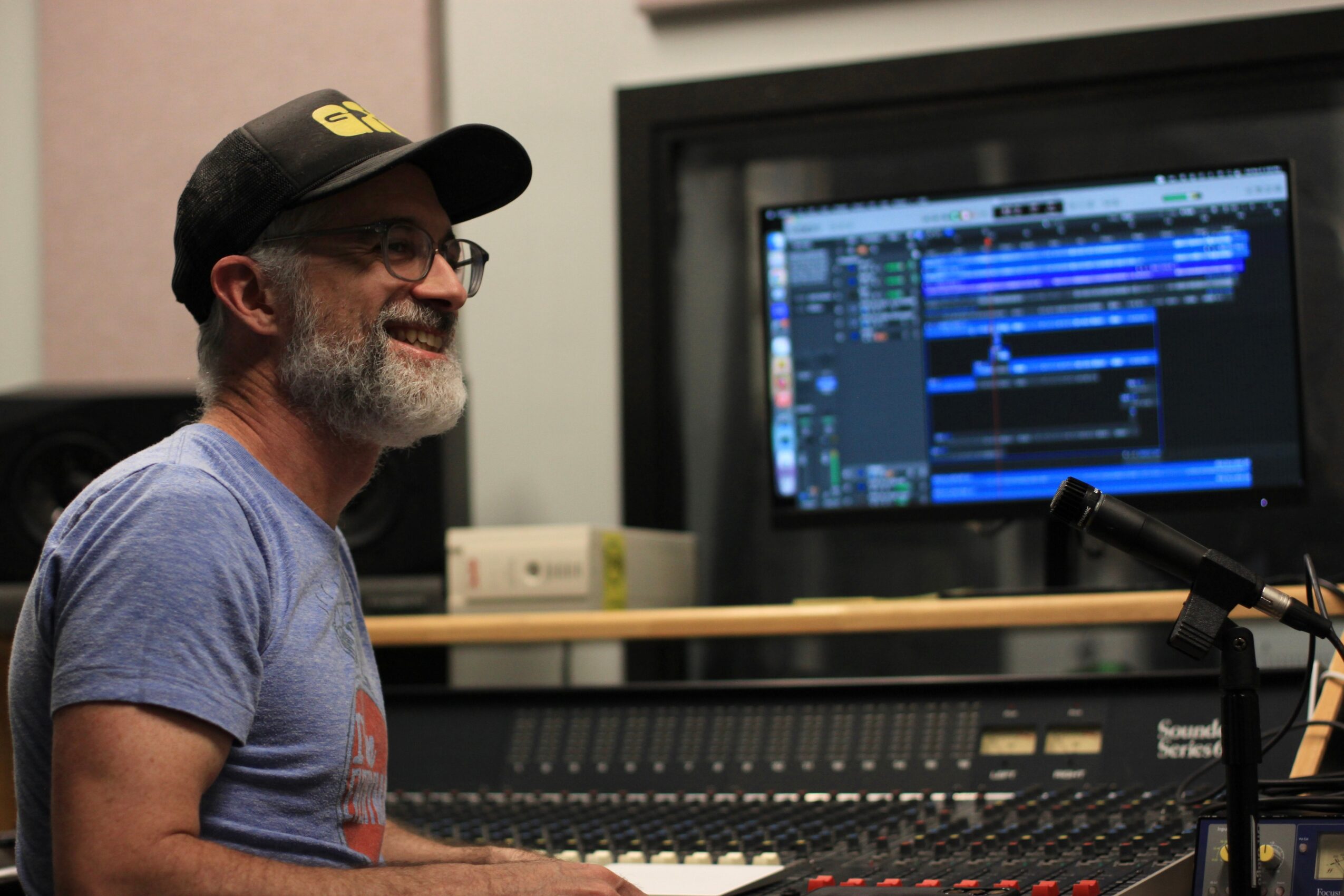 Man with glasses and a beard smiling, sitting in front of a computer screen displaying music editing software.