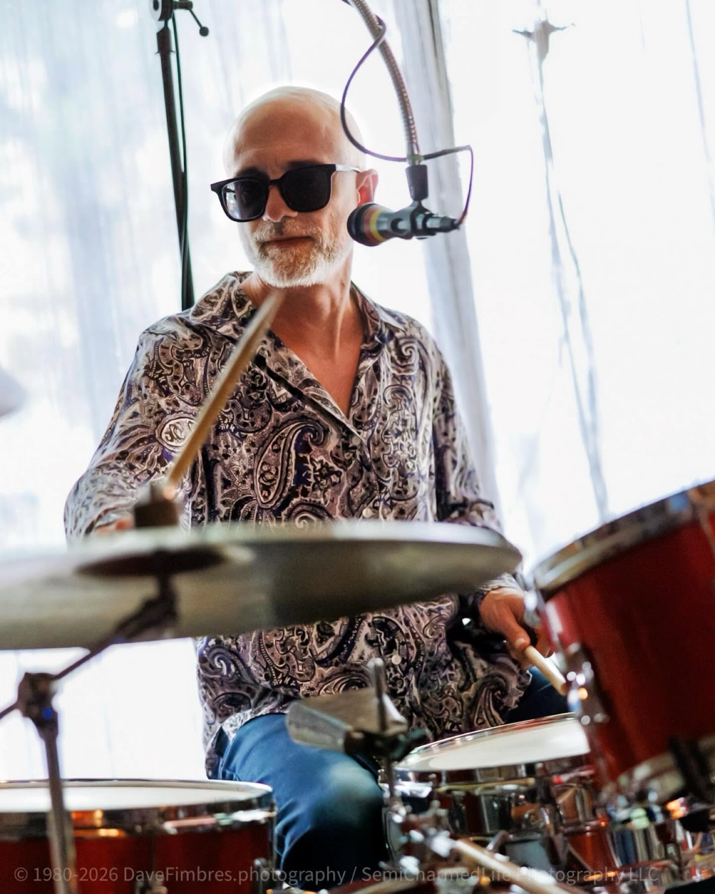 Bald man with sunglasses playing drums in a bright room with curtains, wearing a patterned shirt.