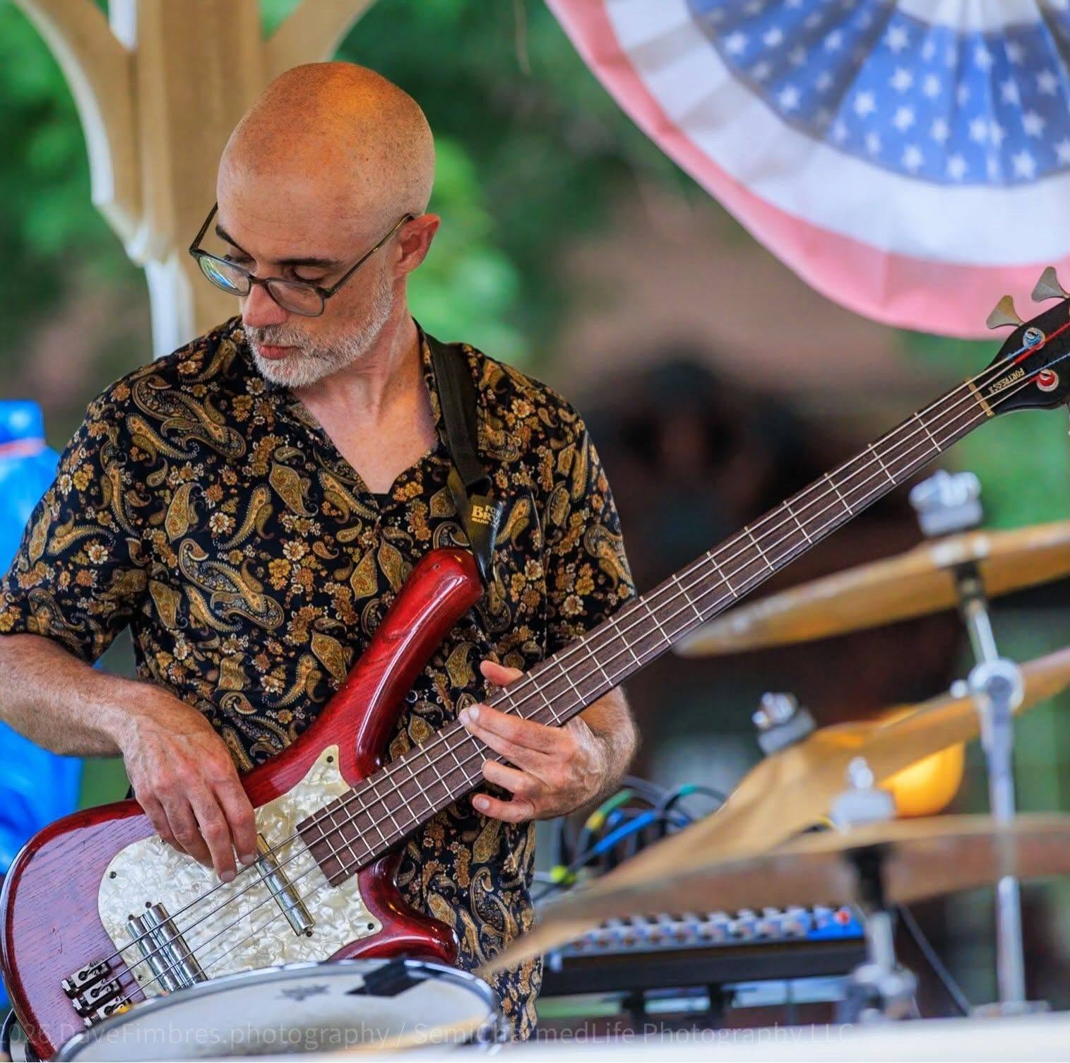 Bald man with glasses playing a red bass guitar outdoors, wearing a patterned shirt, with a blurred background and patriotic decoration.