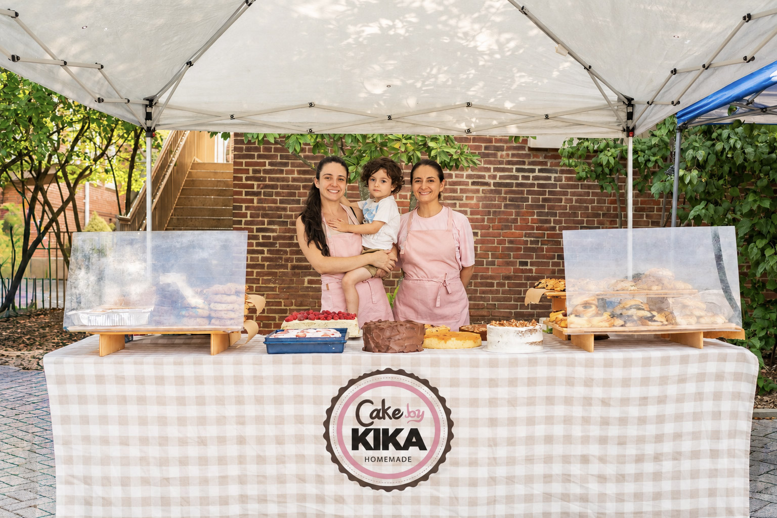 Three people, two women and a child, stand behind a cake stand at an outdoor event, with a brick wall and trees in background.