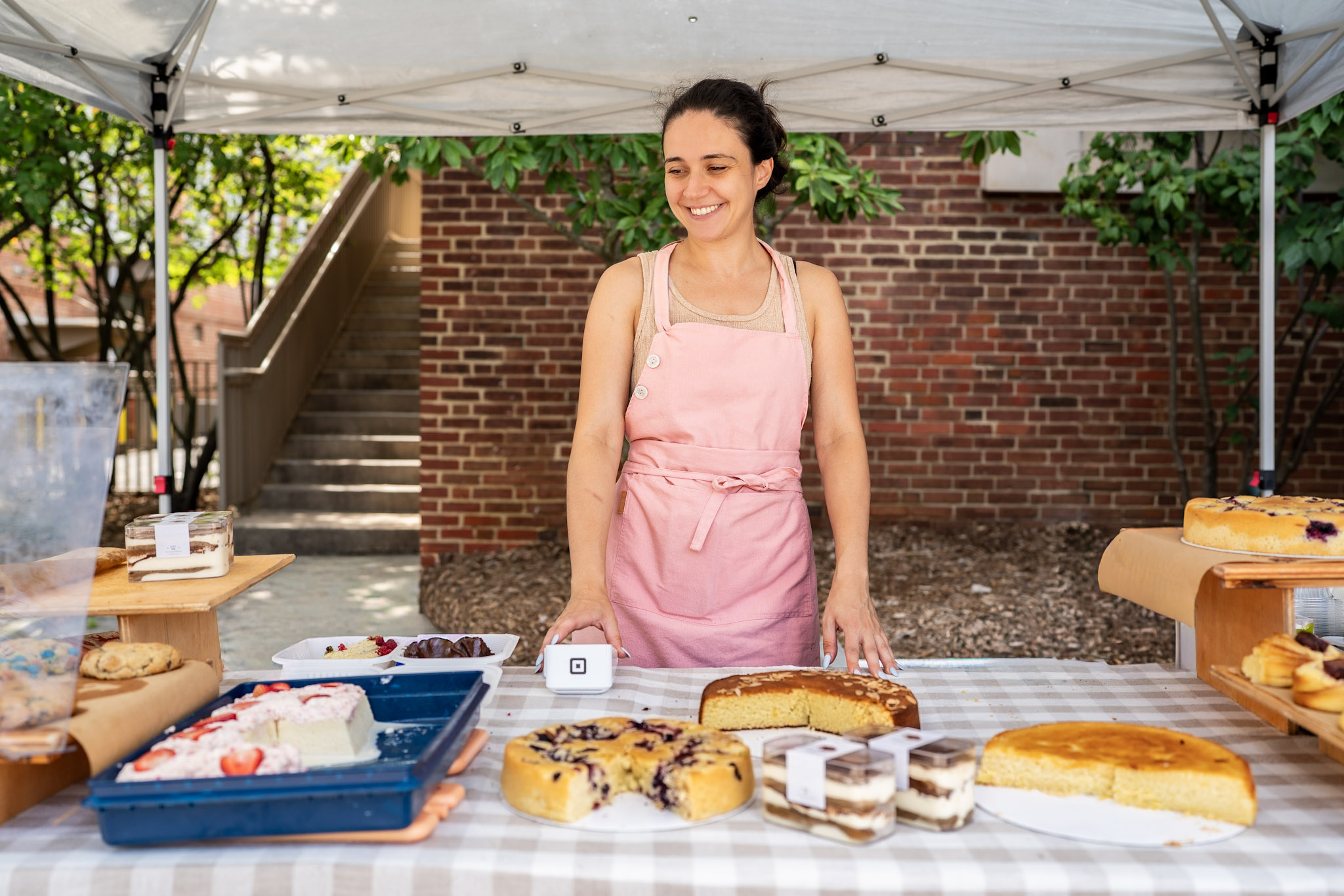 Young woman in pink apron standing behind a table with baked goods outdoors, brick wall and stairs in background.
