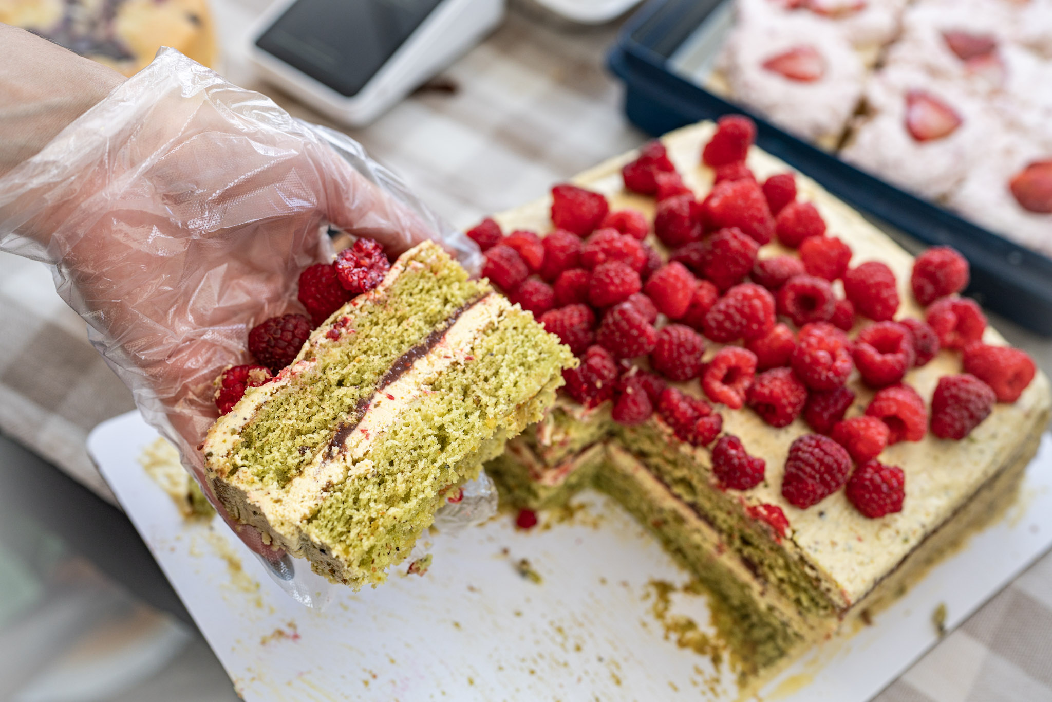 Person in a glove holds a slice of green layered cake with raspberry filling, next to a large raspberry-topped cake on a table.