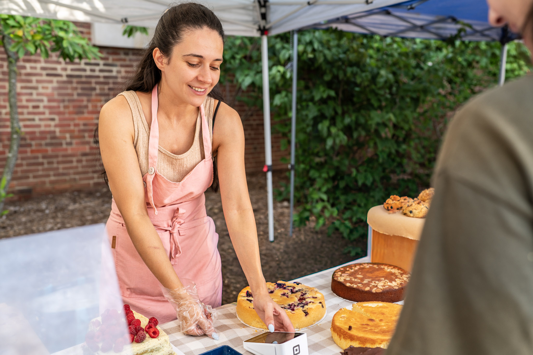 Young woman in pink apron arranging baked goods at outdoor market stall, with trees and brick wall in background.