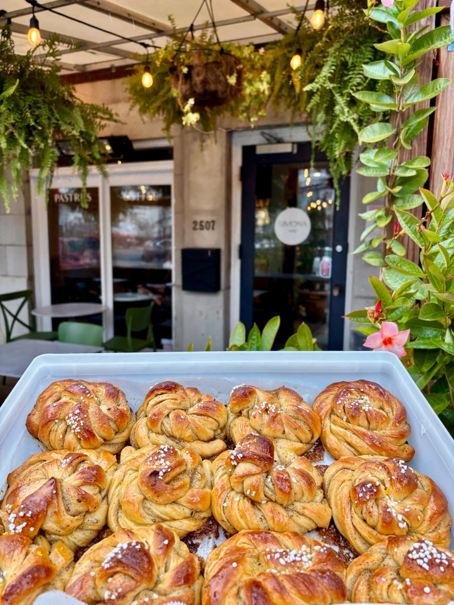 Tray of freshly baked croissants with golden-brown crusts, some topped with sugar crystals, in an outdoor cafe setting with greenery and string lights.