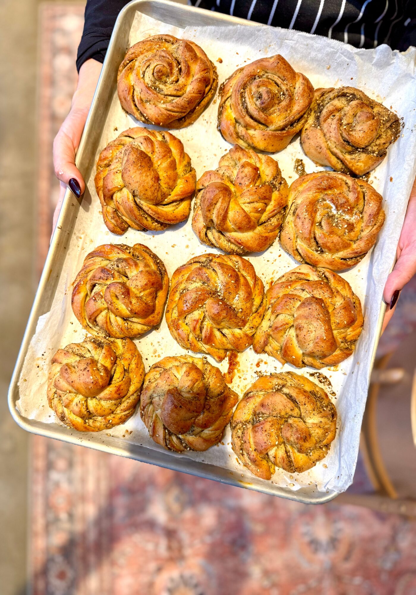 Person holding a tray of freshly baked cinnamon rolls on parchment paper, outdoors, with a blurred background.