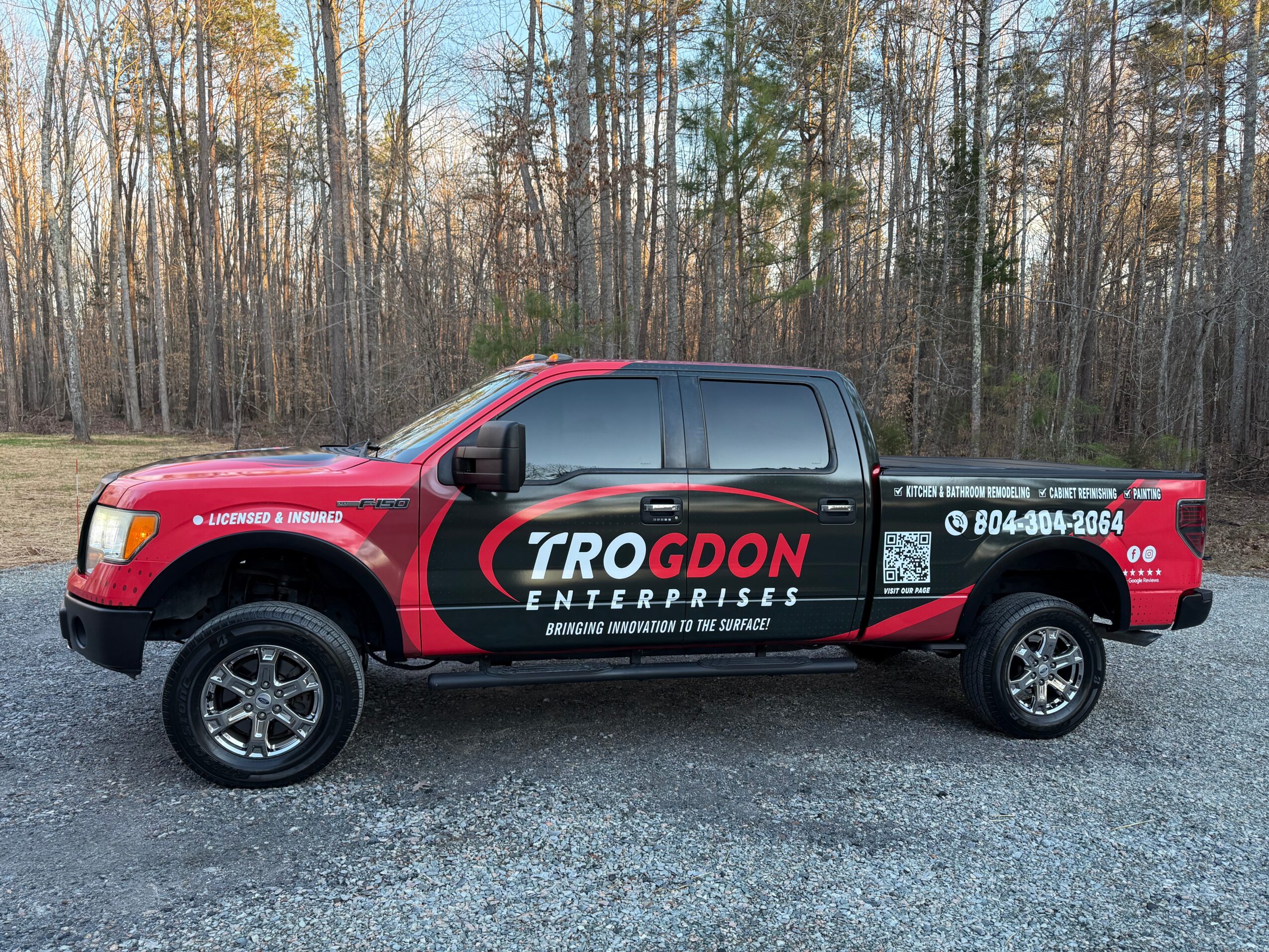 Red and black truck with company logo and contact information parked on gravel, trees in background.
