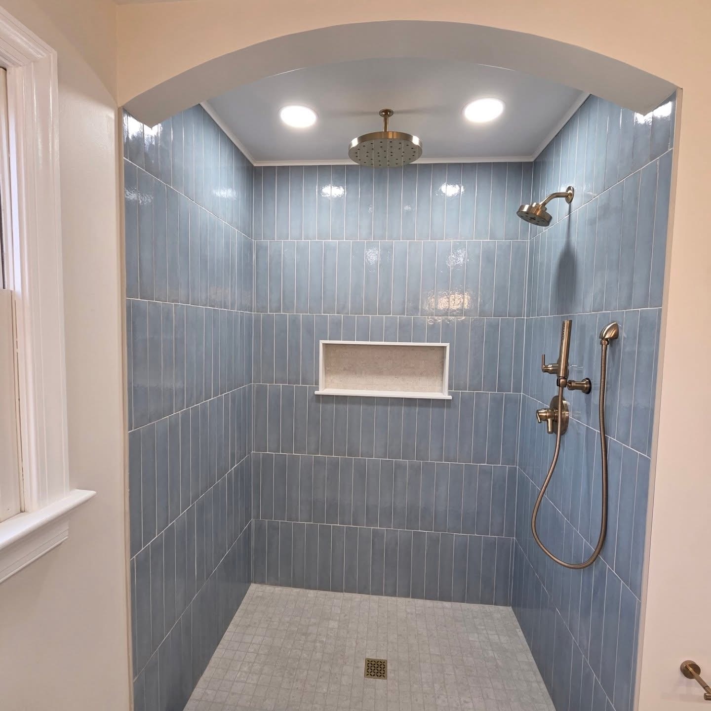 Shower area with blue tiled walls, built-in shelf, ceiling light, and showerhead, viewed through an arched opening.
