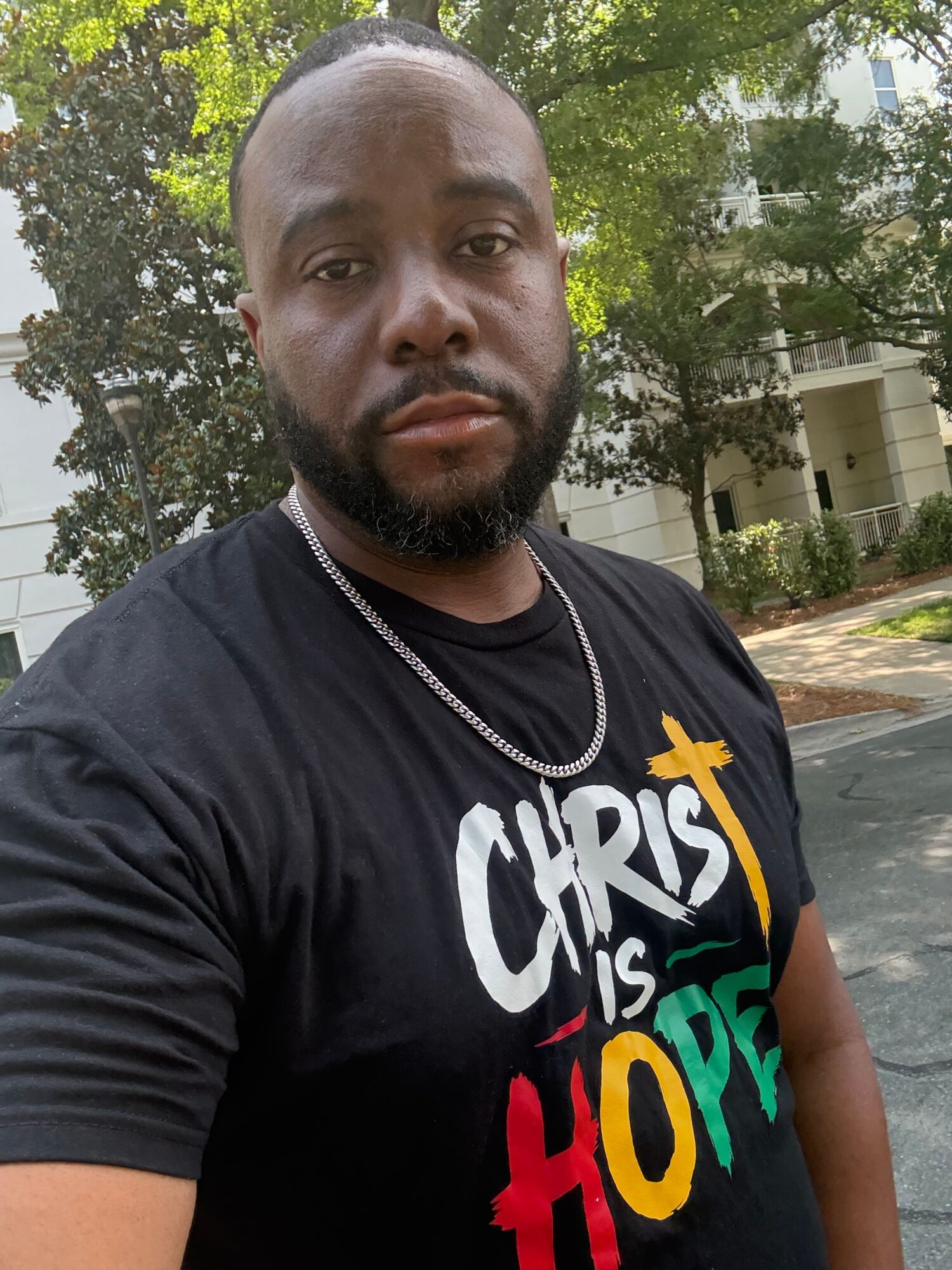 A man with a beard and short hair takes a selfie outdoors, with trees and a building in the background.