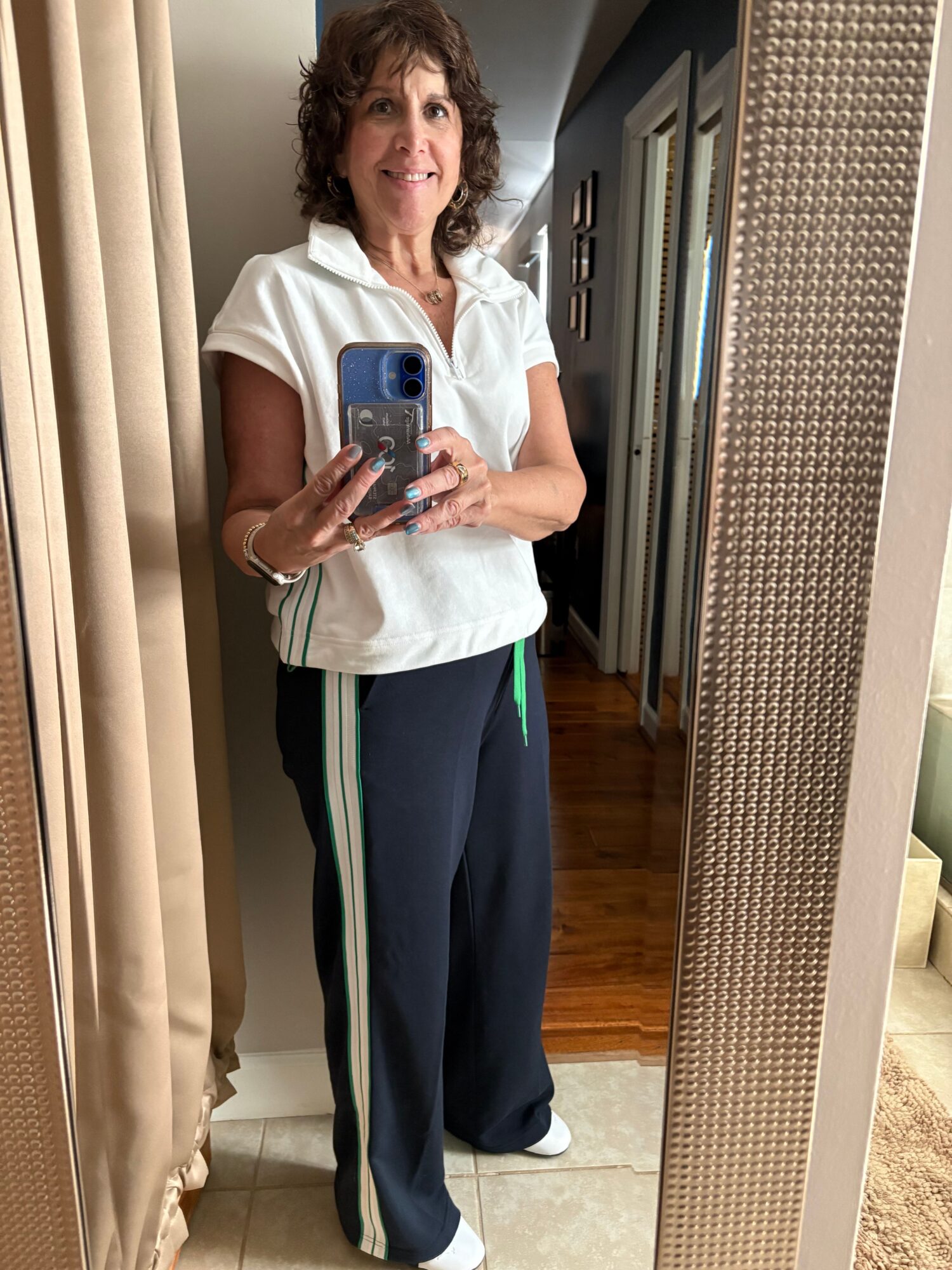 Woman with curly hair taking a mirror selfie in a fitting room, wearing a white top and dark pants with green and white stripes.