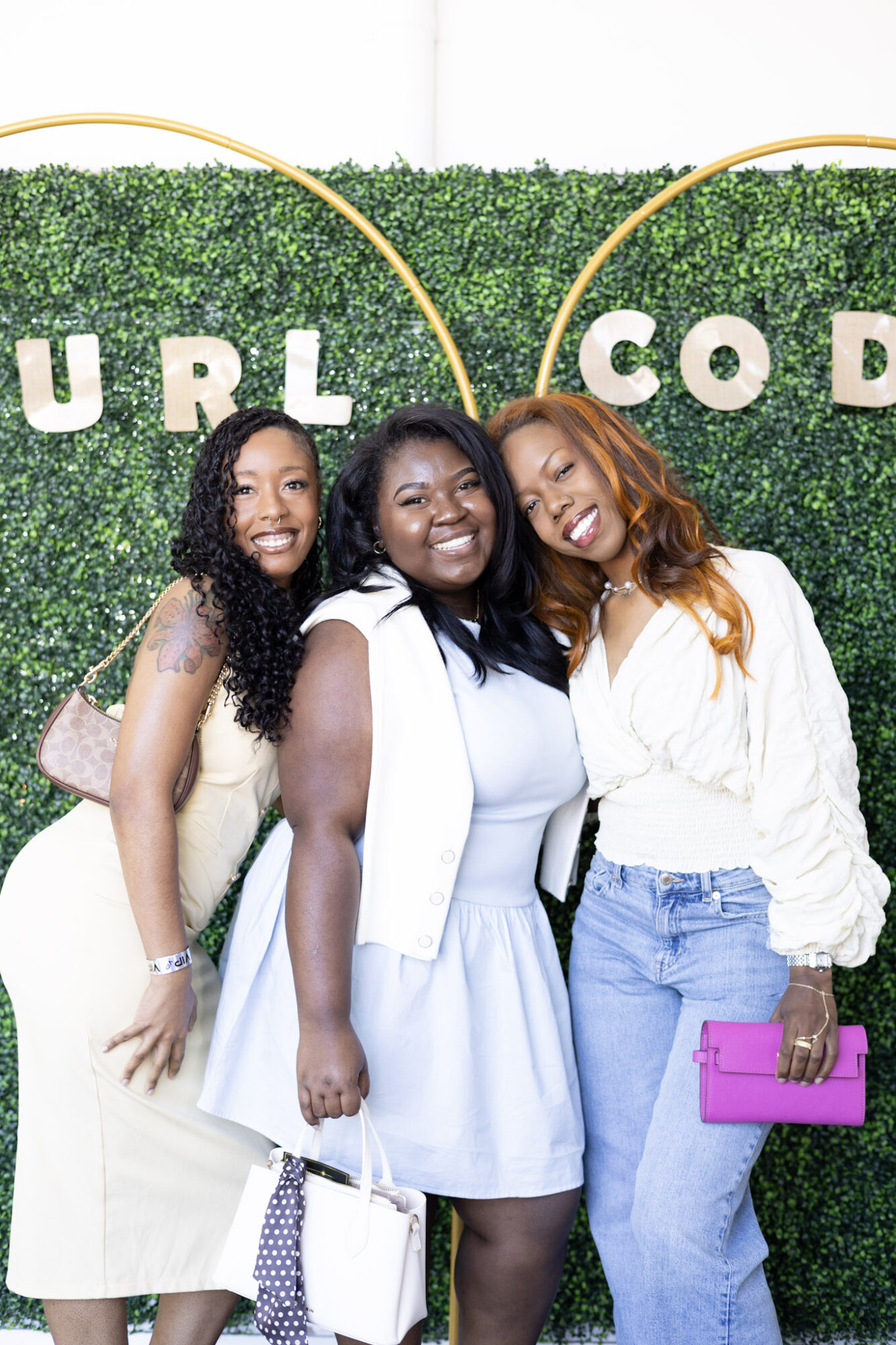 Three women smiling and posing together in front of a green hedge with partial text visible.