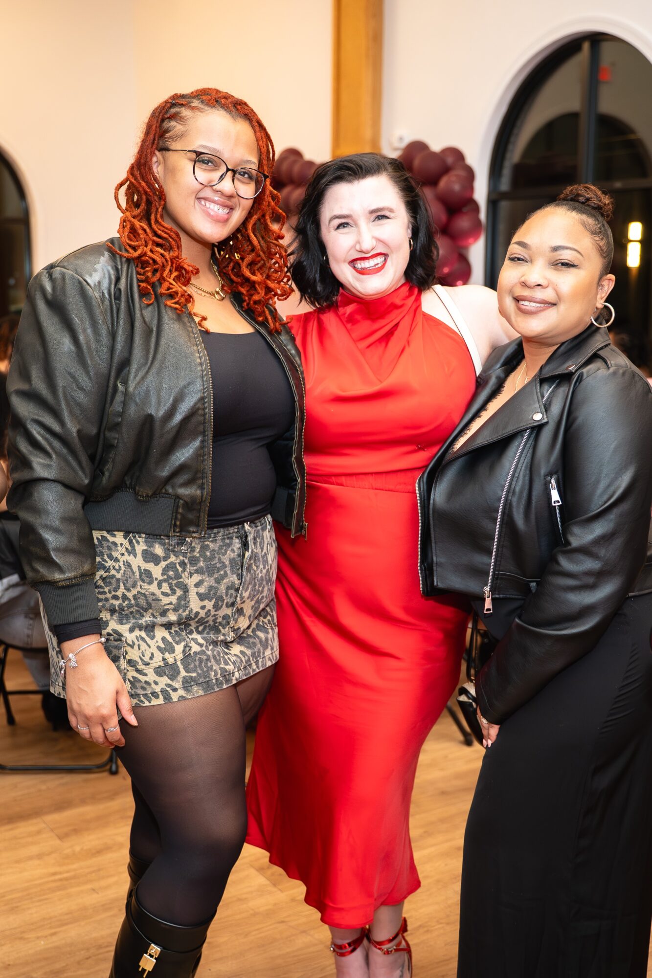 Three women smiling indoors, one in a red dress, others in black jackets, standing close together.