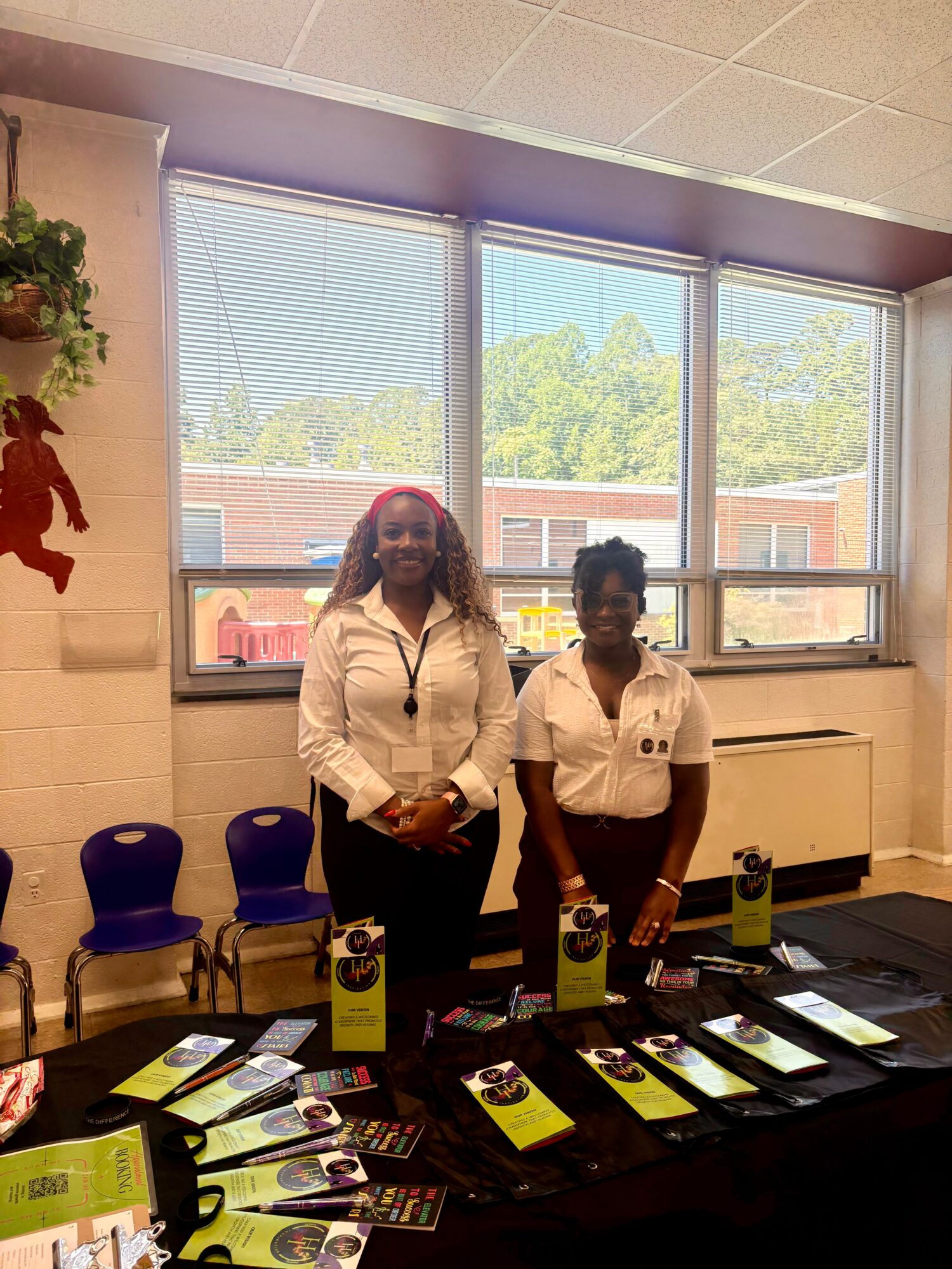 Two women standing behind a table with brochures and promotional materials, in a room with large windows and chairs.