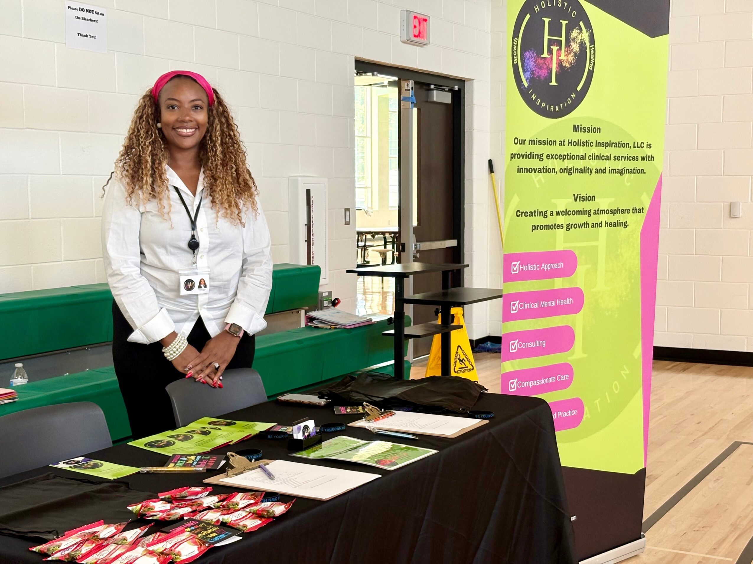 Smiling woman with curly hair and pink headphones standing behind a table with brochures and items, in a gymnasium or event space.