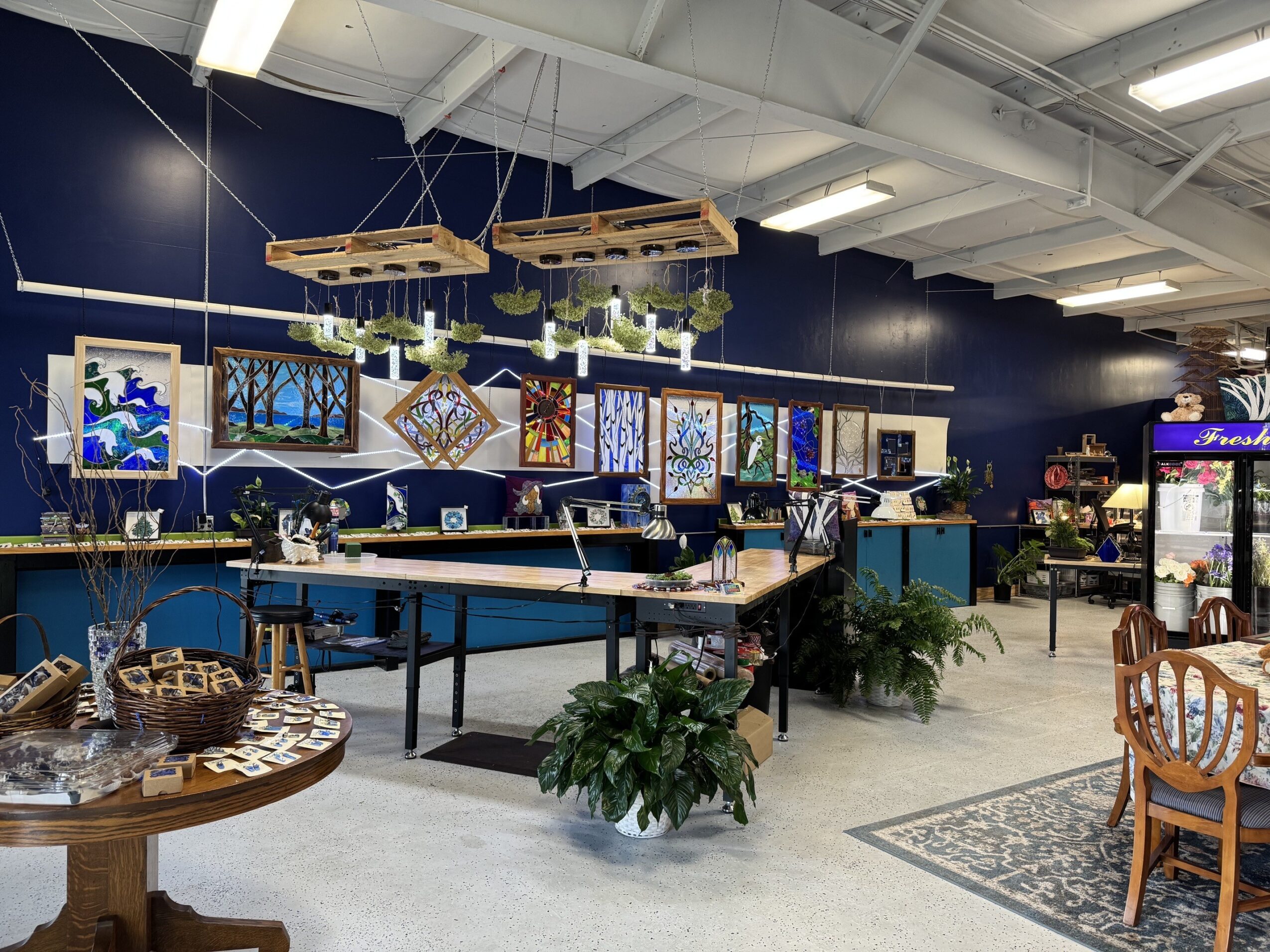 Indoor space with artwork on a dark blue wall, tables, chairs, and plants, resembling an art gallery or showroom.