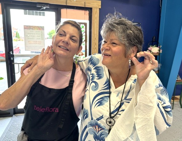 Two women smiling and talking, one wearing a black apron and the other with gray hair and a patterned blouse.