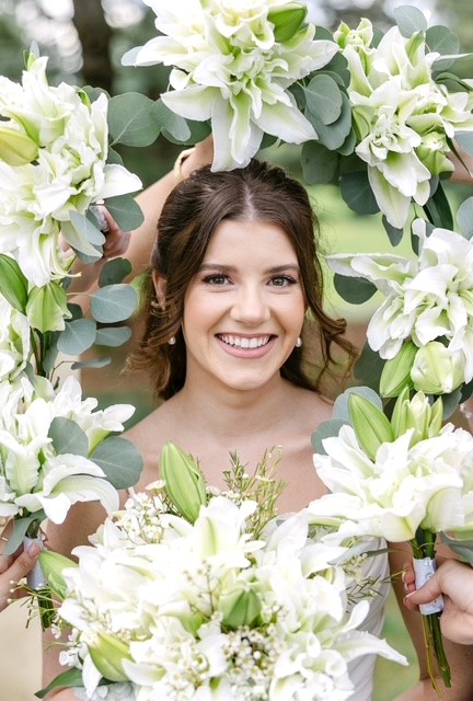Smiling woman surrounded by white flowers and green leaves, holding a bouquet, with floral arch framing her face.