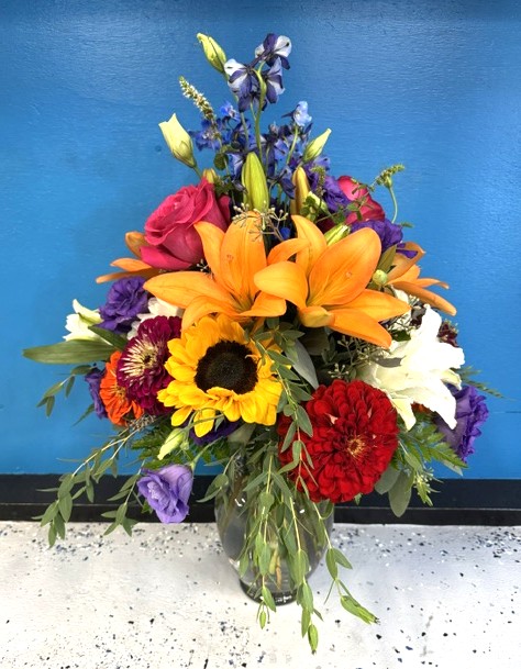 Colorful flower bouquet with lilies, sunflowers, roses, and other flowers in a glass vase against a blue background.