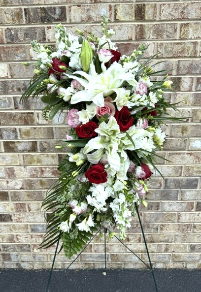 Flower arrangement with white lilies, roses, and greenery on a stand against a brick wall