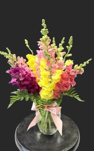 Colorful flower bouquet with pink, yellow, purple, and green flowers in a glass vase with a pink ribbon, on a dark surface.