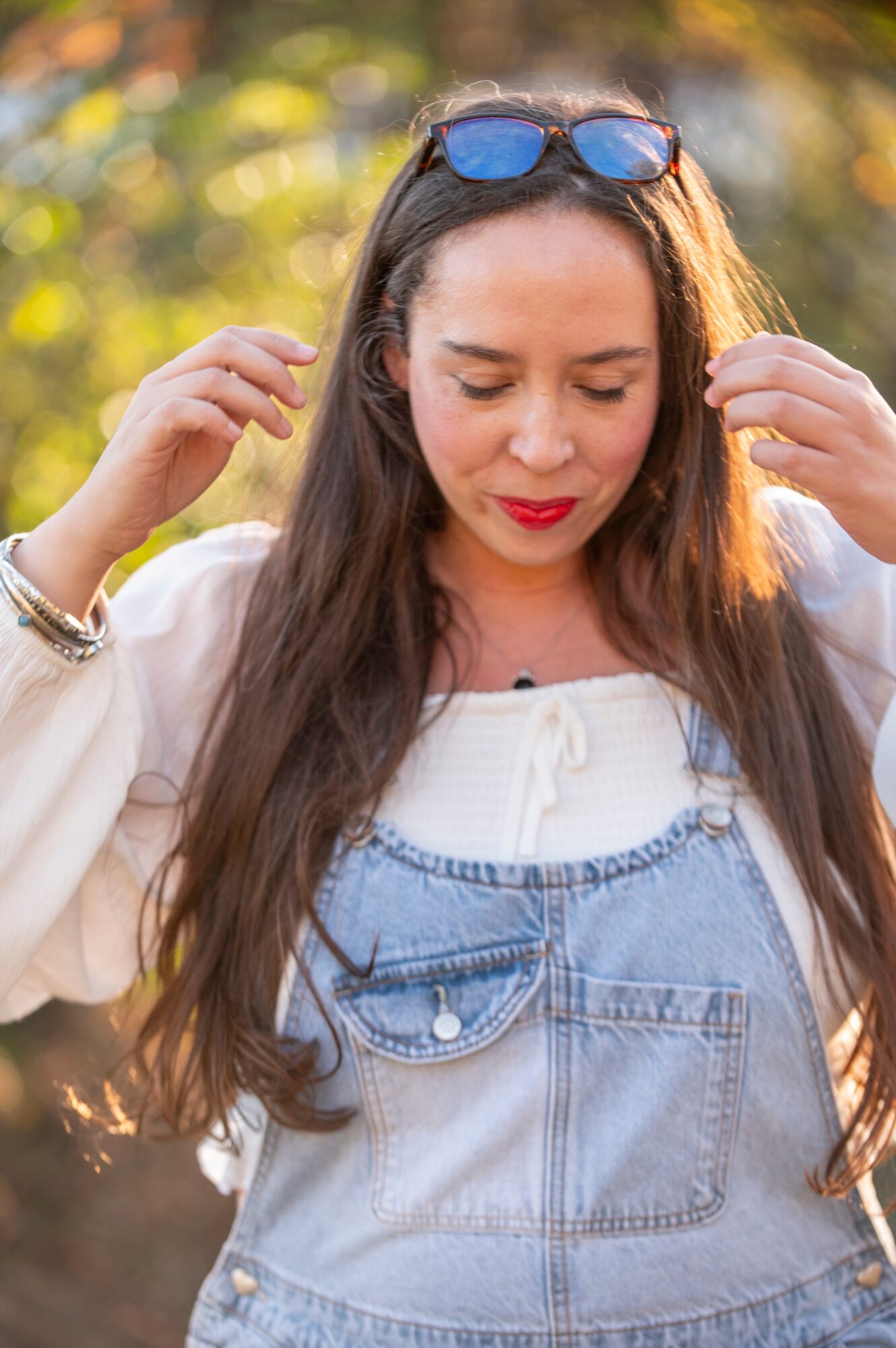 Young woman with long hair, sunglasses on her head, wearing a white top and denim overalls outdoors.