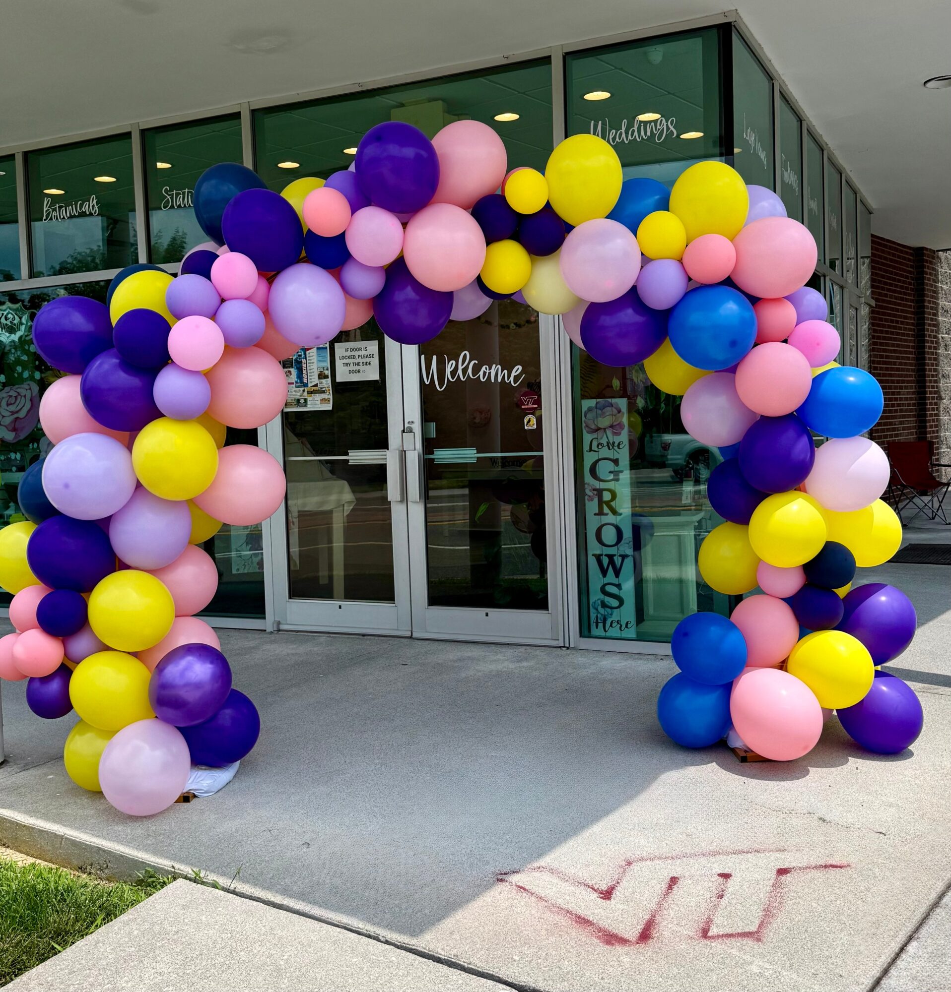Balloon arch with purple, pink, yellow, and blue balloons in front of glass doors, decorated for a welcoming event.