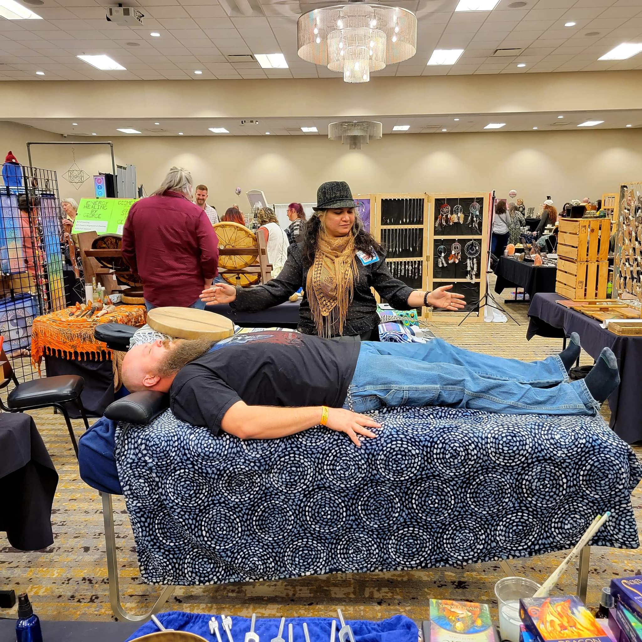 Person lying on a table at a market stall, with a woman standing behind, surrounded by various items and other shoppers.