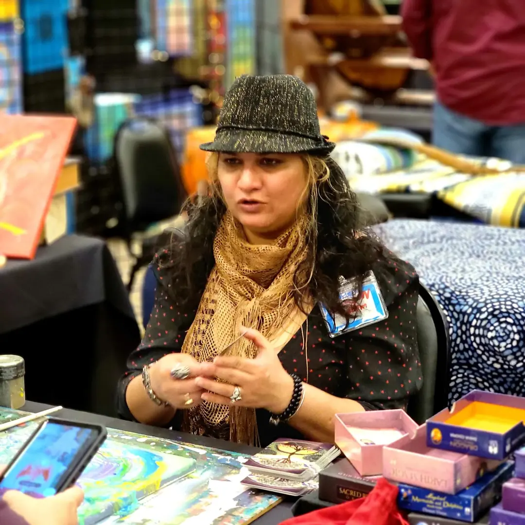 Woman wearing a hat and scarf sitting at a table with various items, in a busy indoor setting.