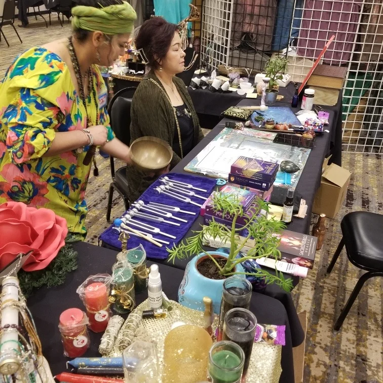 Two women browsing a table with jewelry, plants, and decorative items at an indoor market or craft fair.