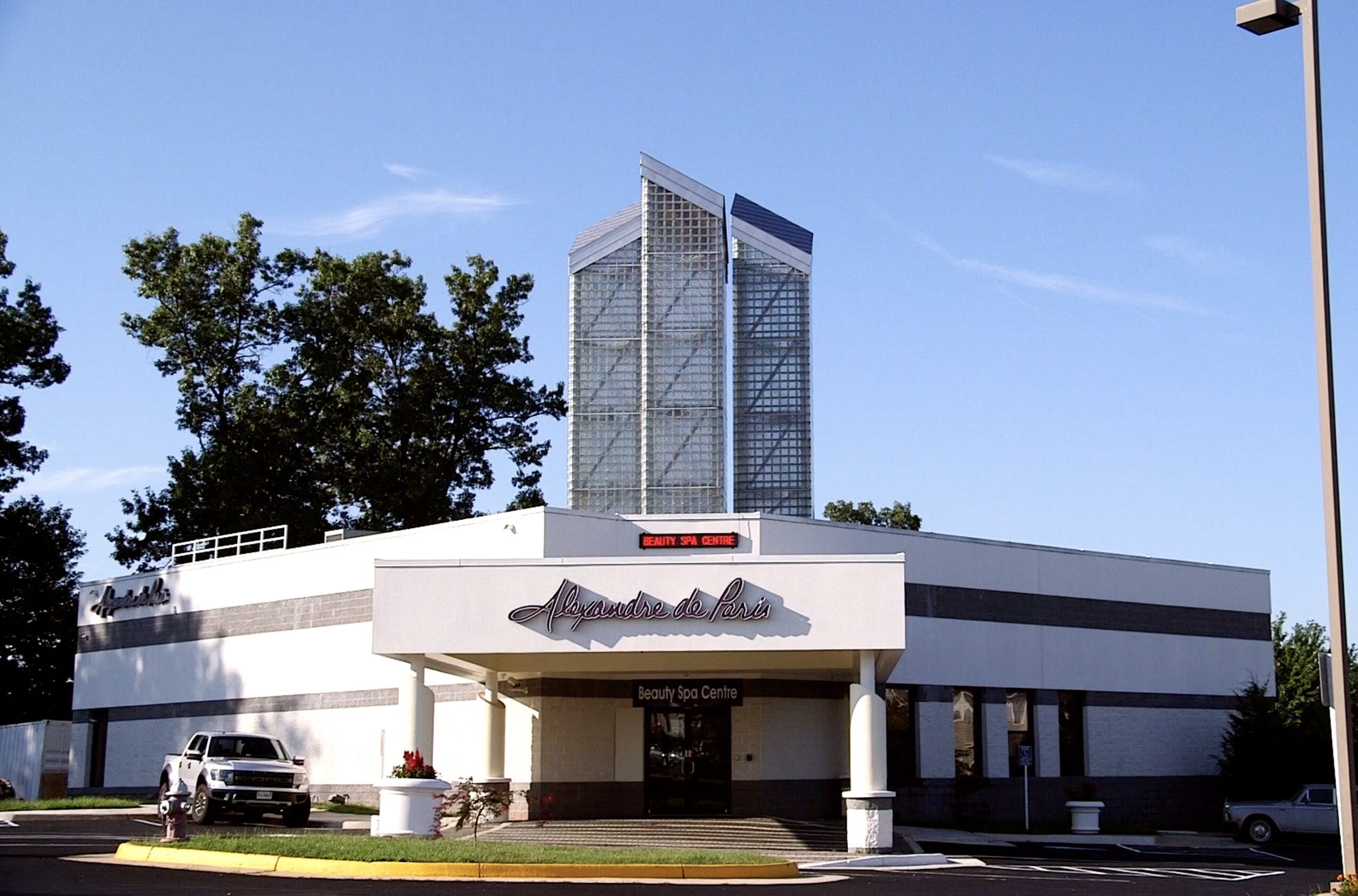 Modern building with a tall glass tower in the background, trees, and a parking area in front.