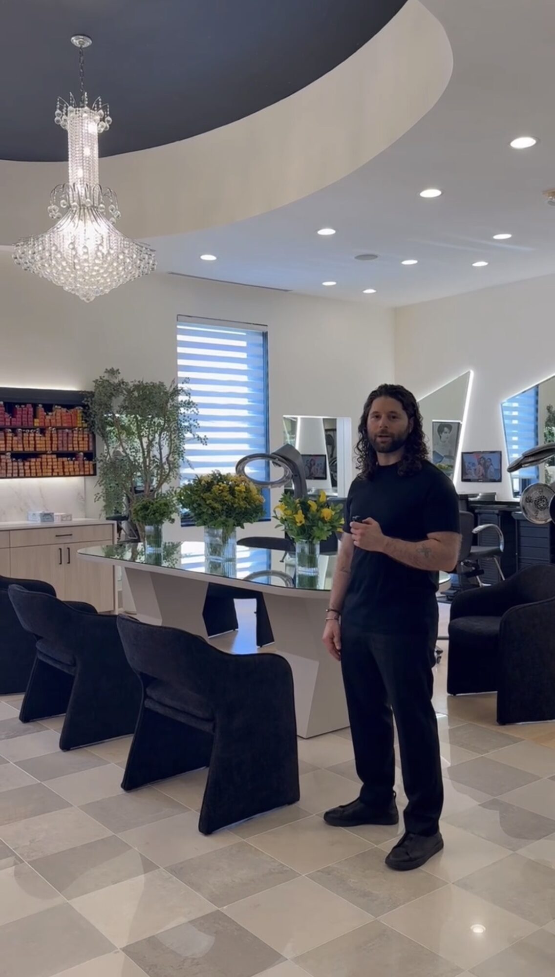 Man standing in a modern, well-lit room with a chandelier, plants, and black chairs, holding a phone or object.