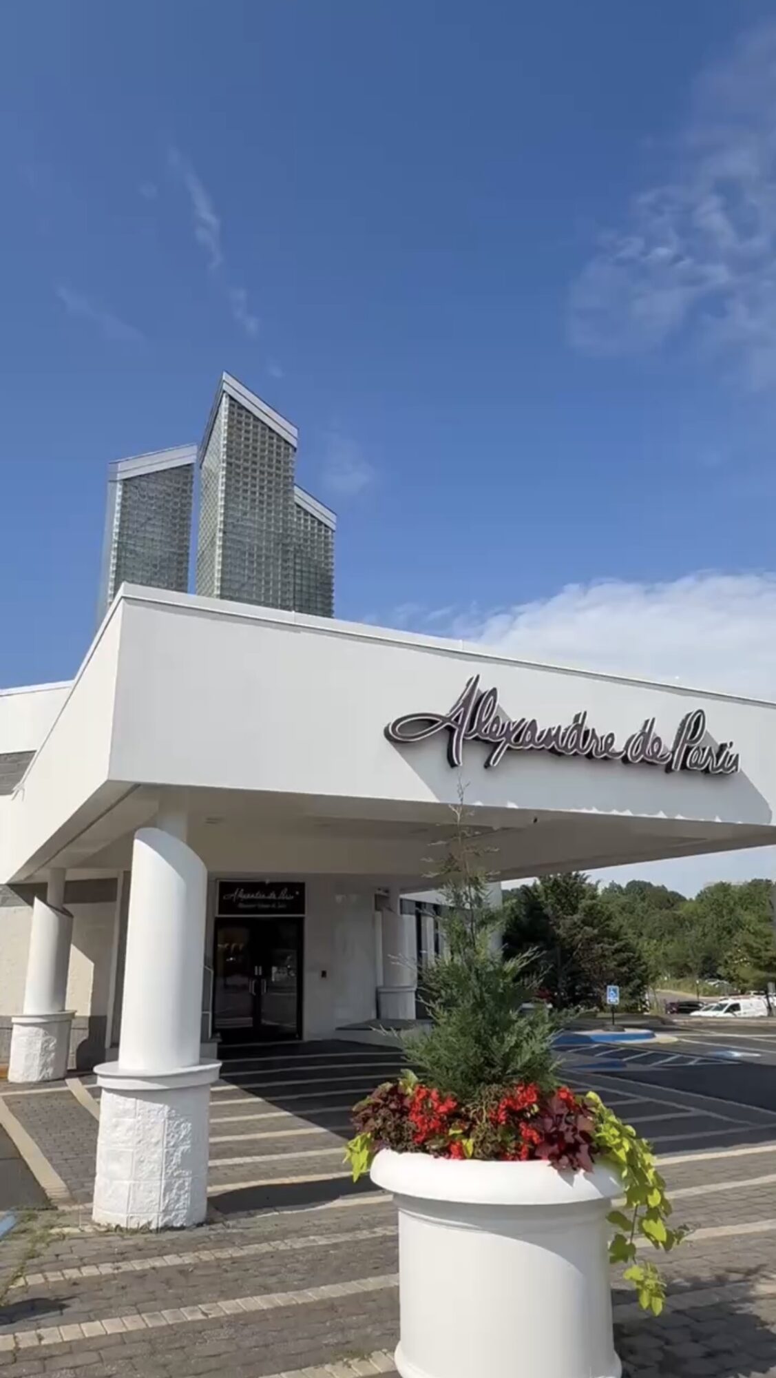 Building entrance with a sign, white columns, and a flower planter, under a blue sky with clouds.