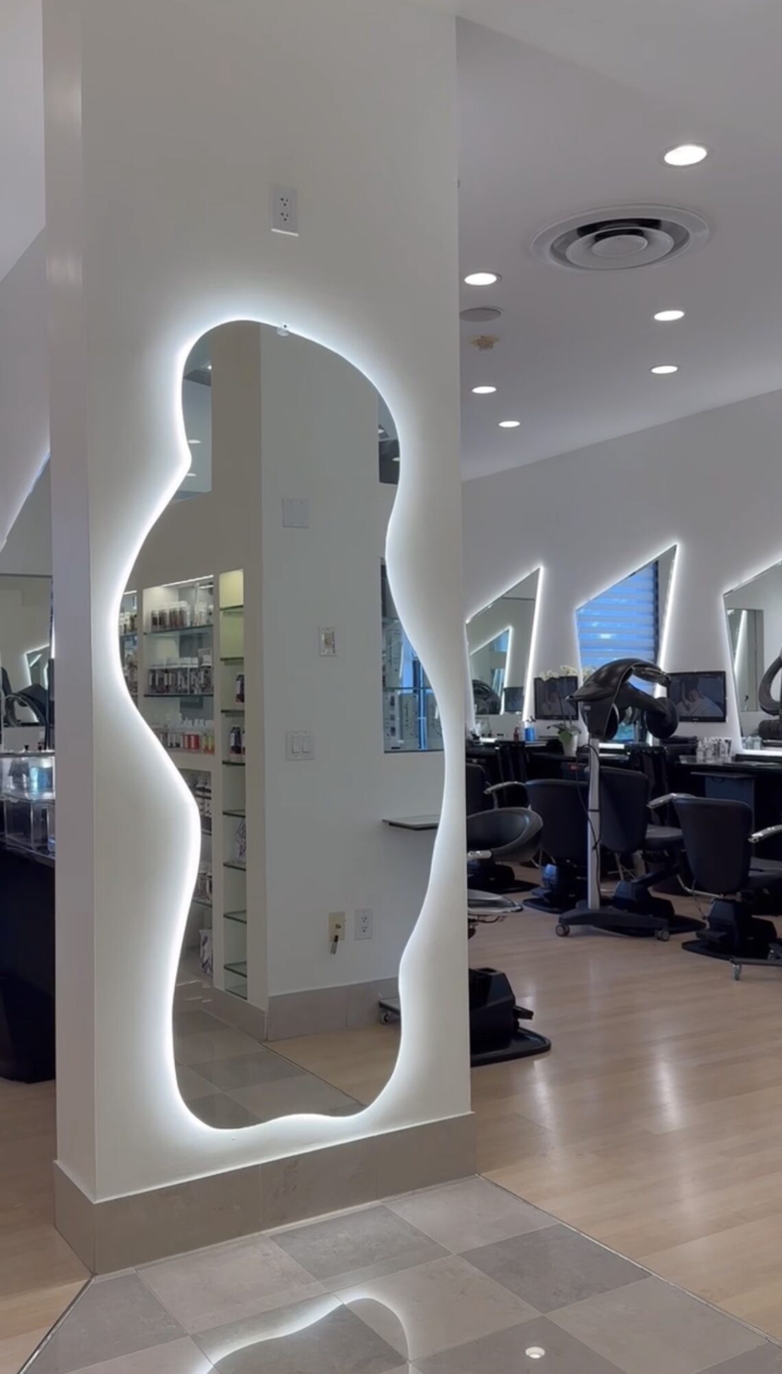 Full-length mirror with irregular, glowing frame in a modern office space with desks and computers.