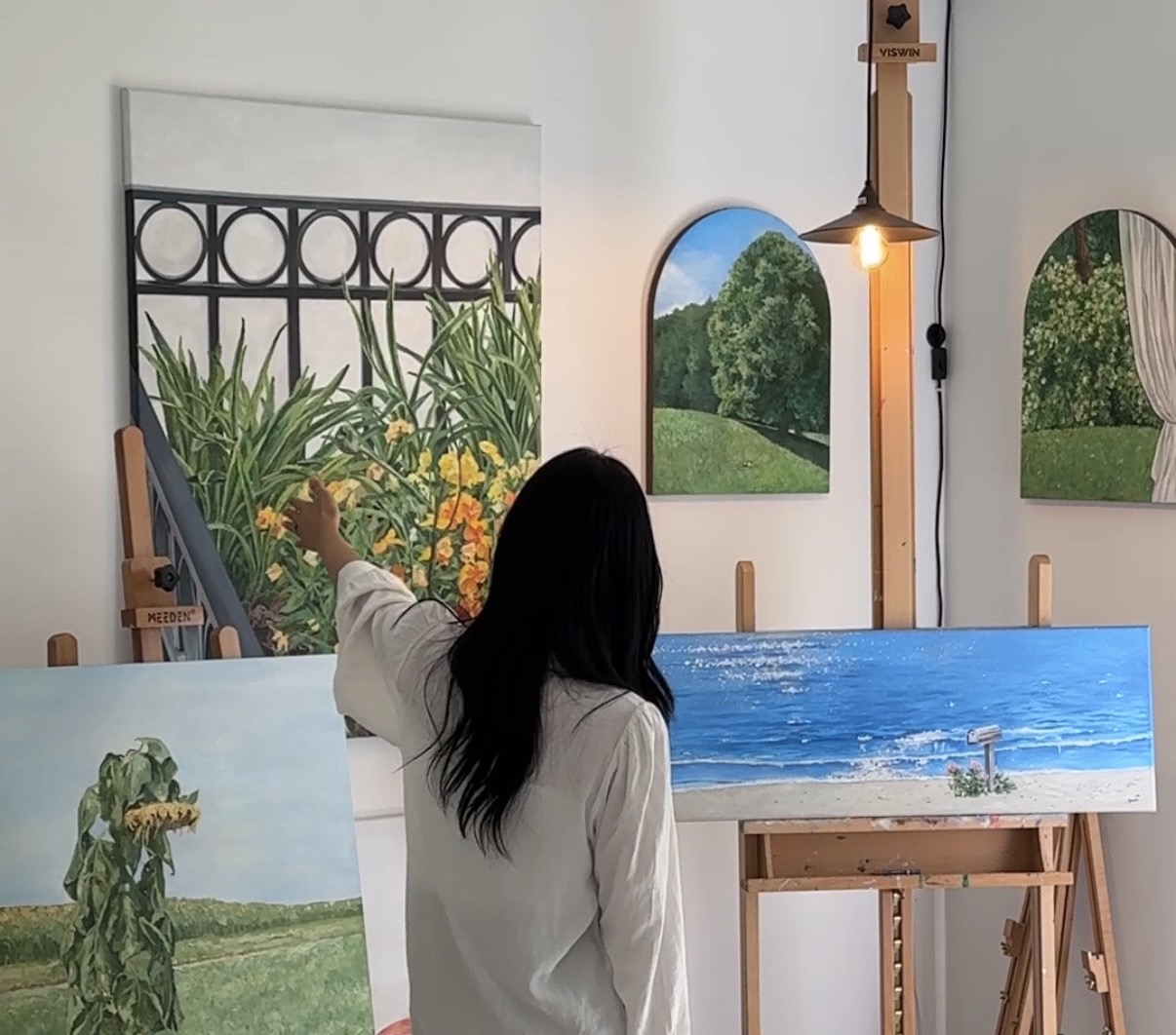 Person with long dark hair in white shirt pointing at artwork in art studio, surrounded by paintings and easels.