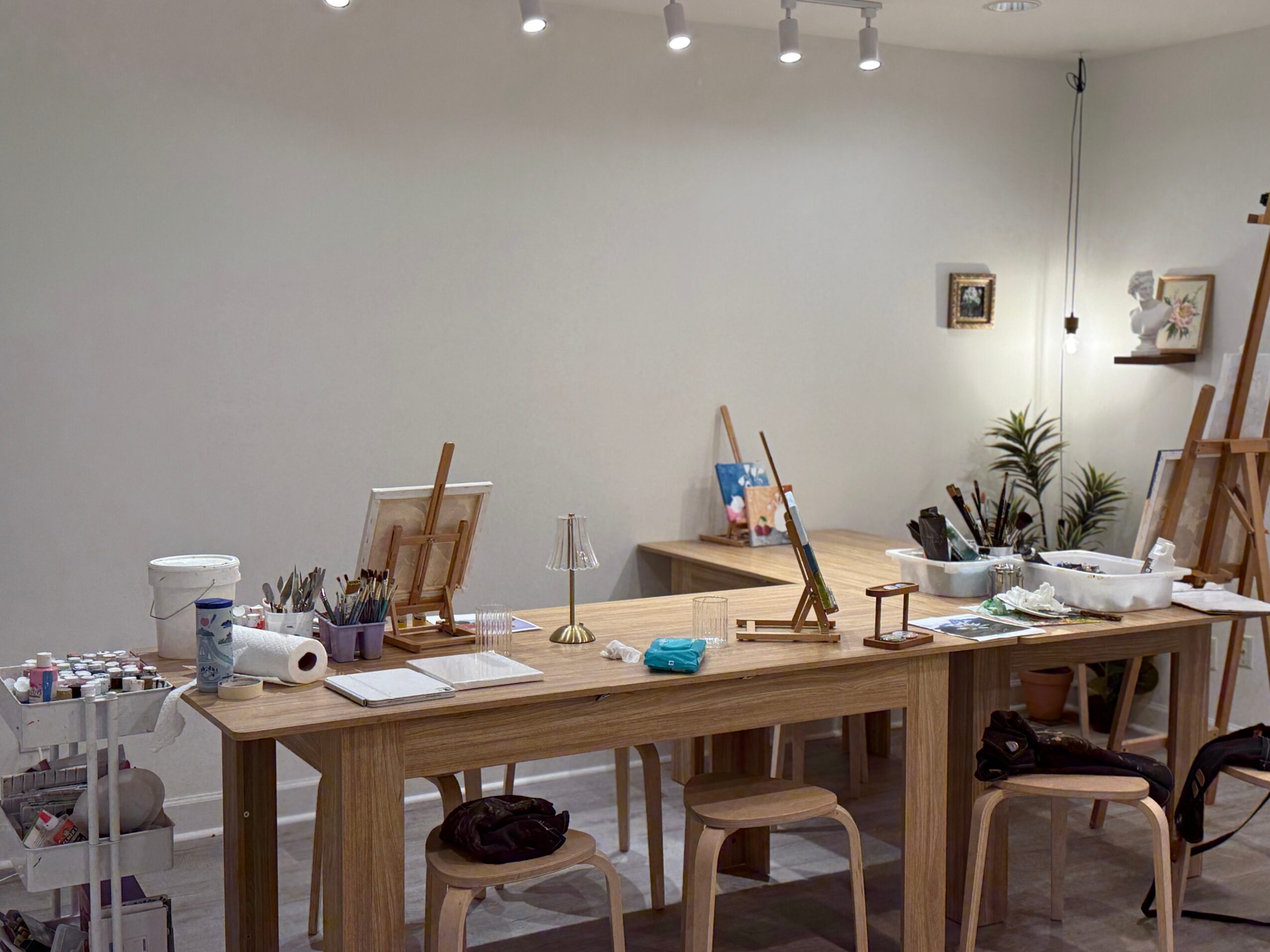 Long wooden table with art supplies, easels, and chairs in a bright room with white walls and small wall decorations.