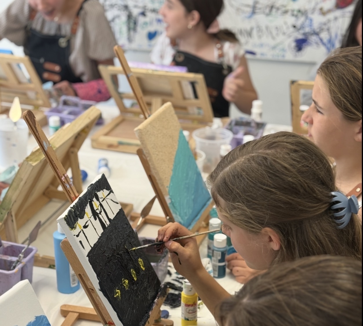 Children painting on canvases at a table, with easels and art supplies visible.