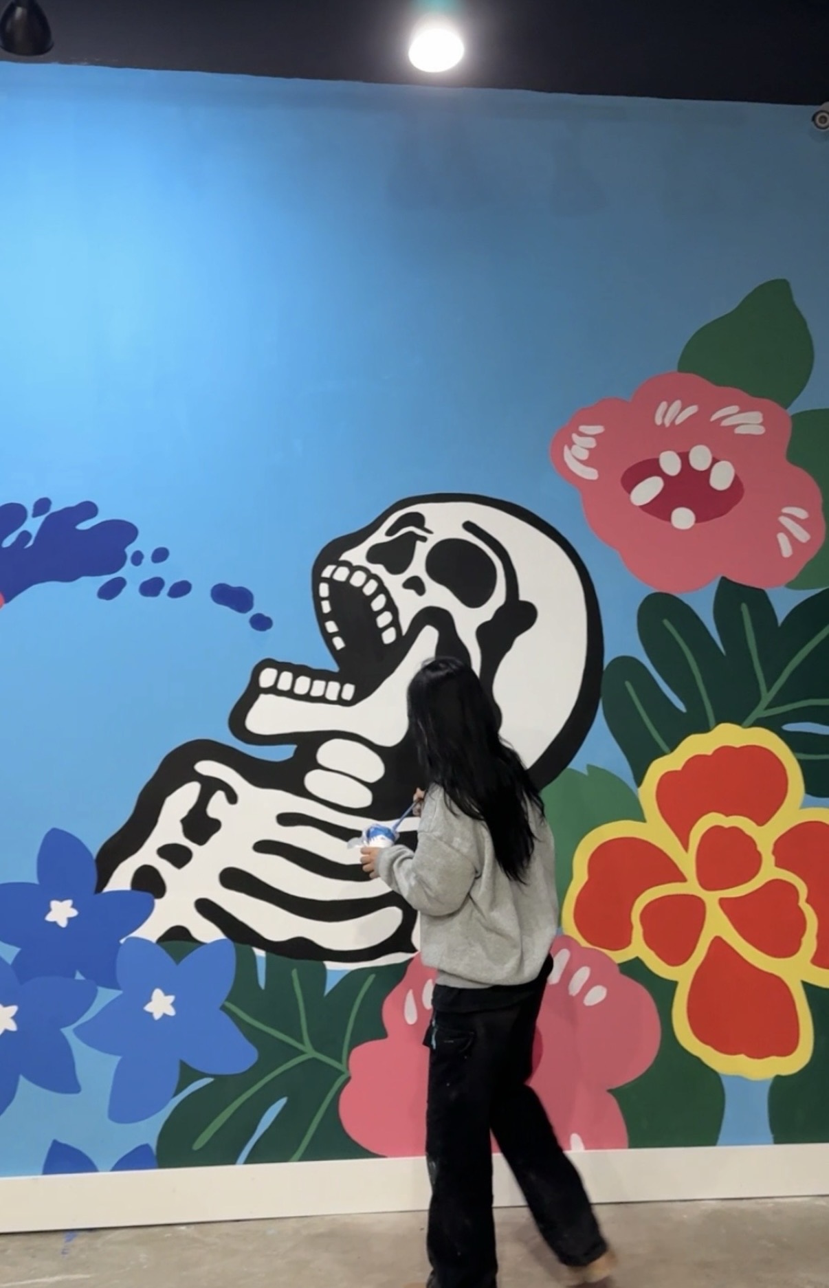 Person with long dark hair standing in front of a colorful mural featuring a skull and vibrant flowers.
