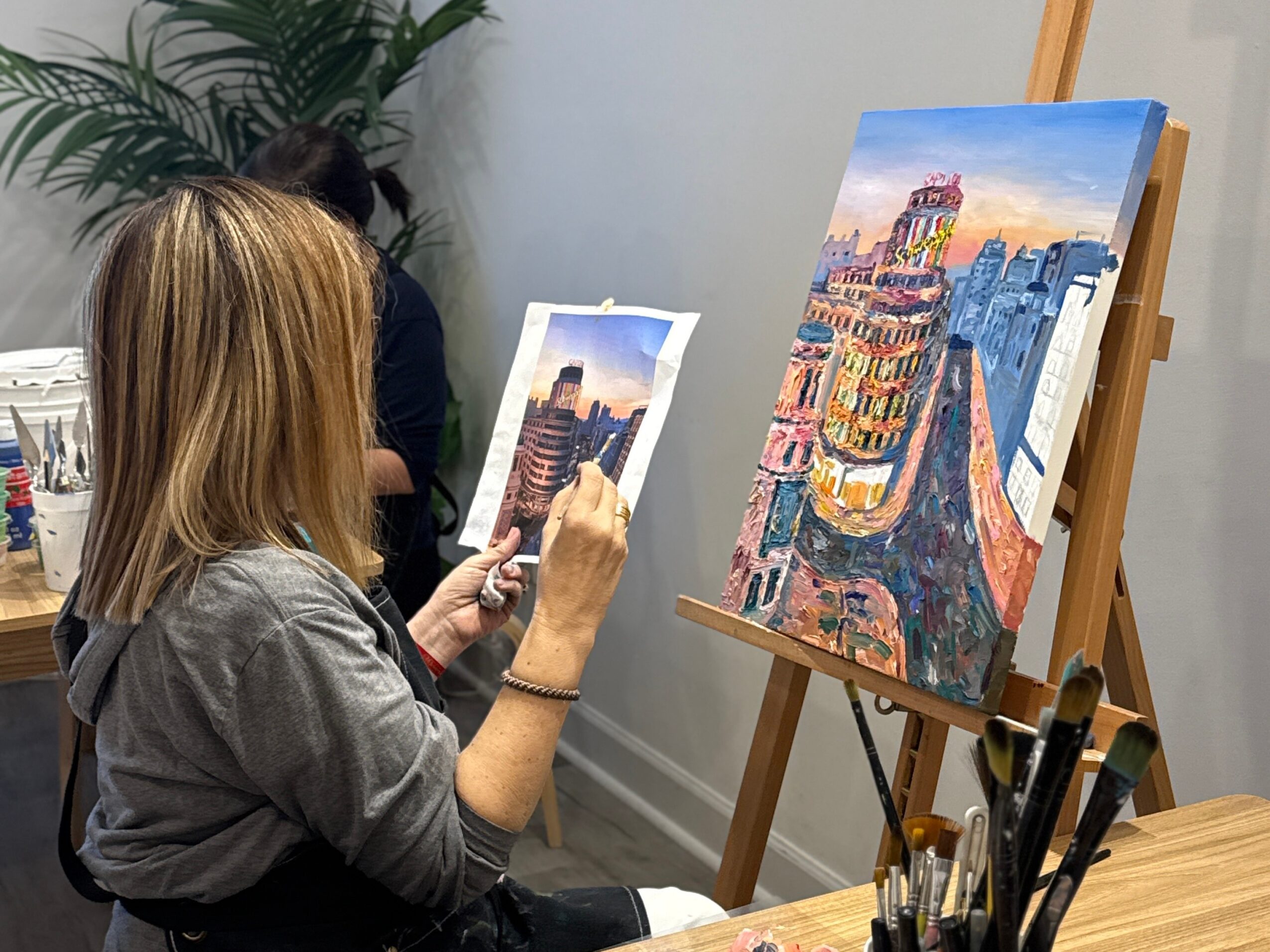Woman with blonde hair painting a cityscape on canvas, with another painting of a city scene on an easel nearby, in an indoor setting.