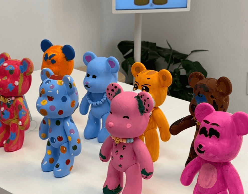 Colorful bear-shaped figurines with various patterns and colors on a white surface, with a monitor and plant in the background.