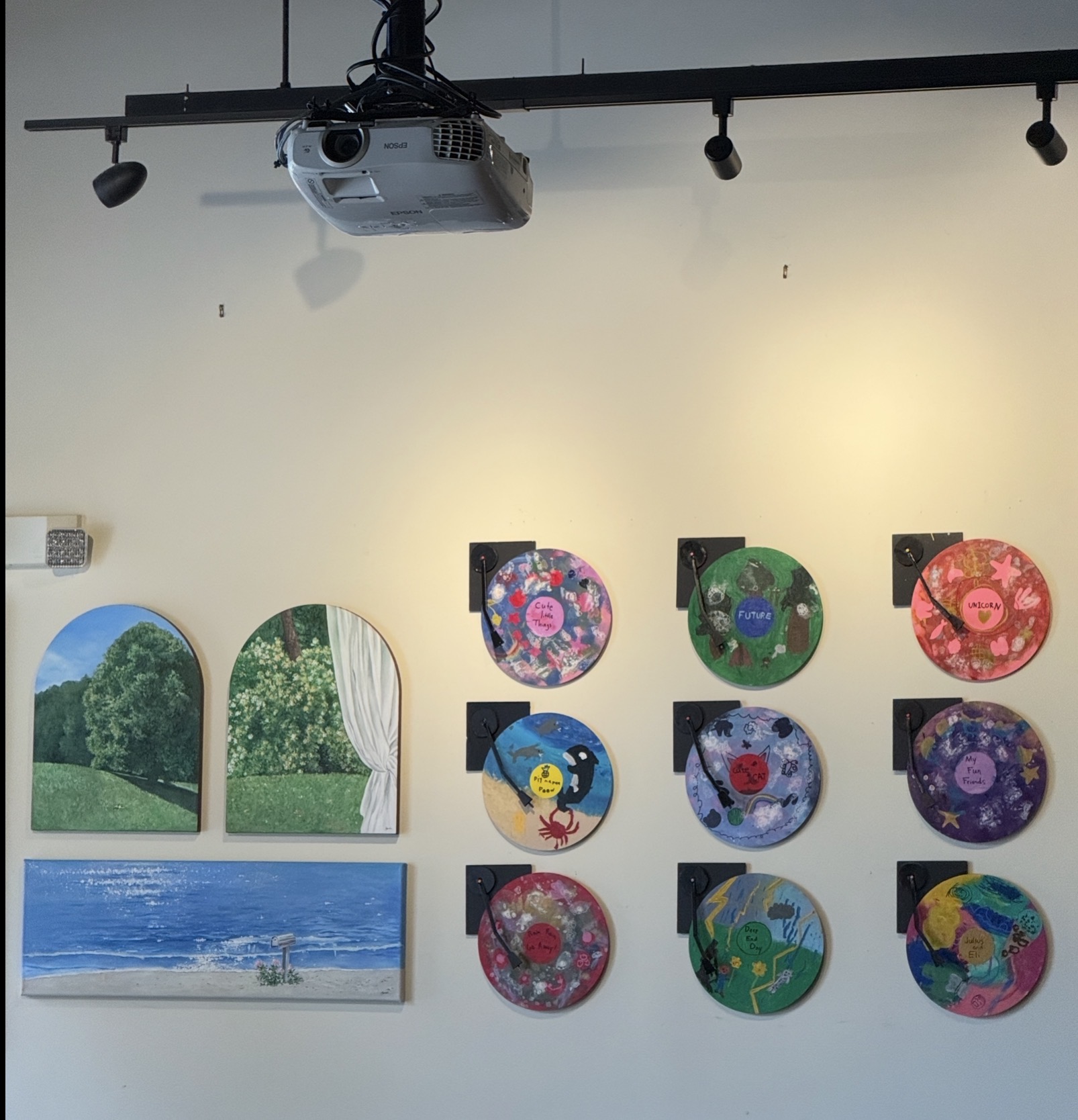 Gallery wall with nine circular and two rectangular art pieces, a projector, and track lighting on a white wall.
