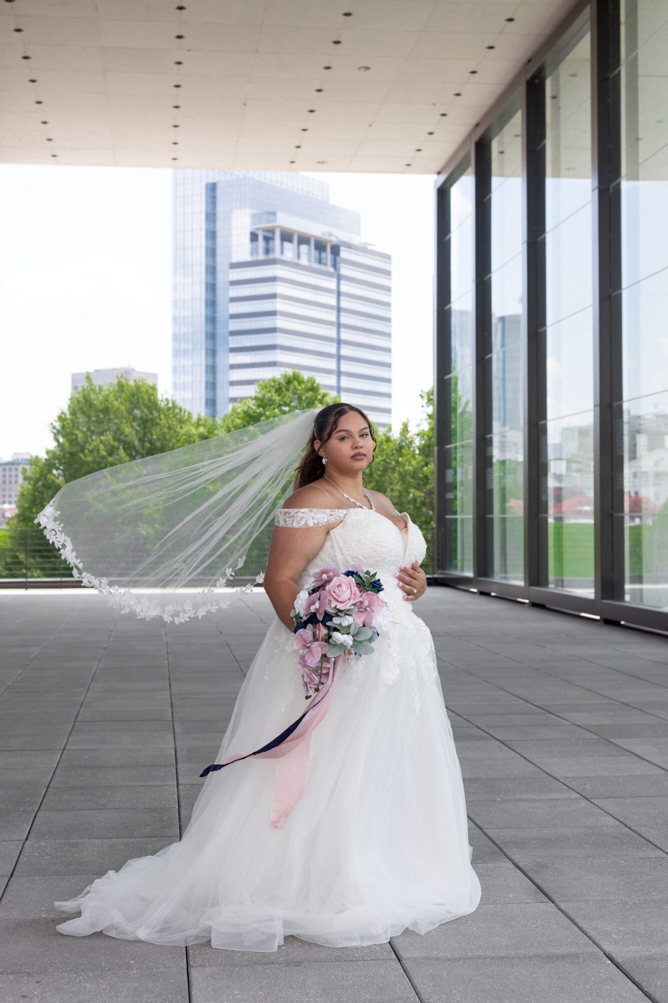 Bride in wedding dress holding bouquet, standing indoors near large glass windows with cityscape view behind her.