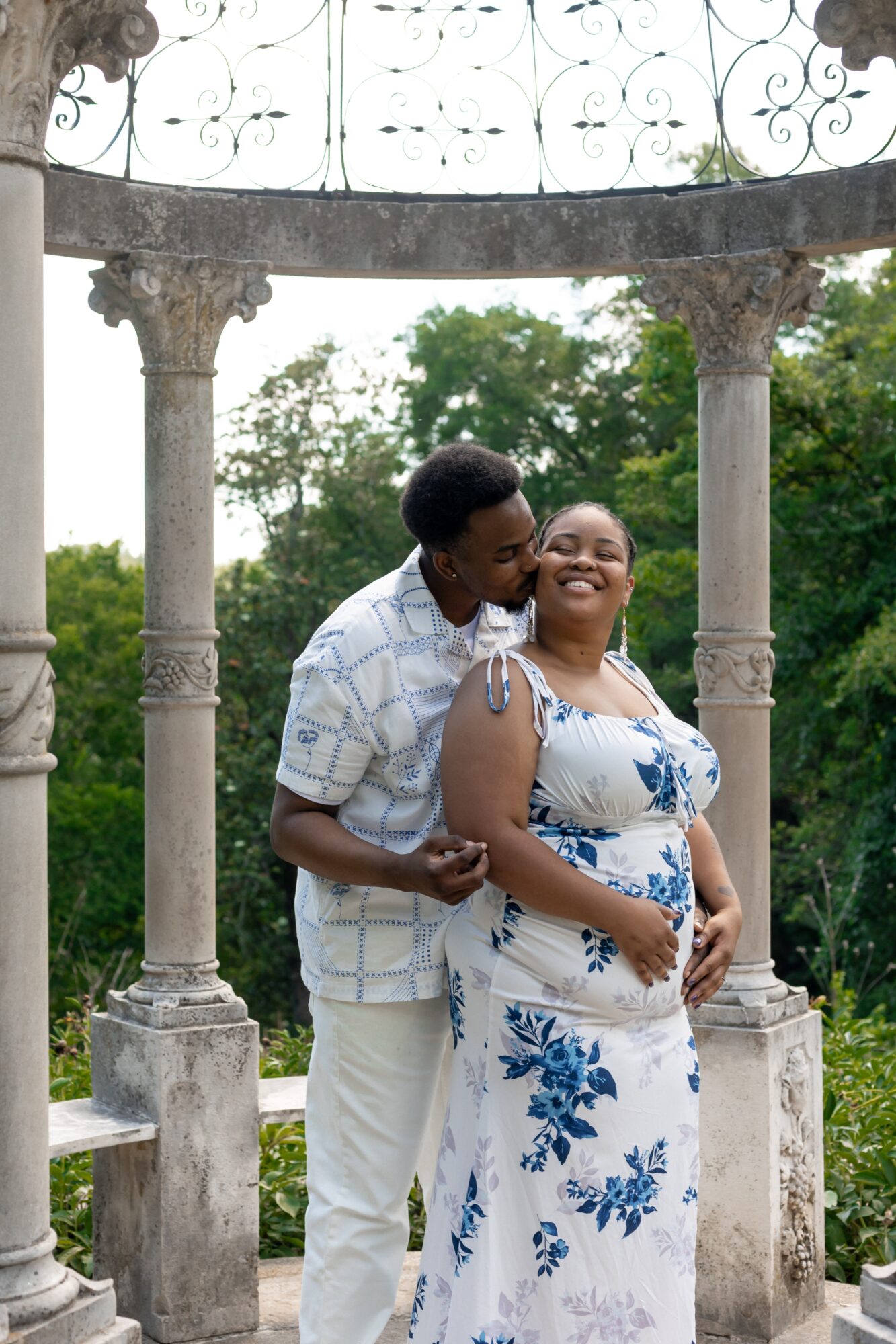 A couple stands under a stone gazebo with columns, smiling and embracing outdoors with trees in the background.