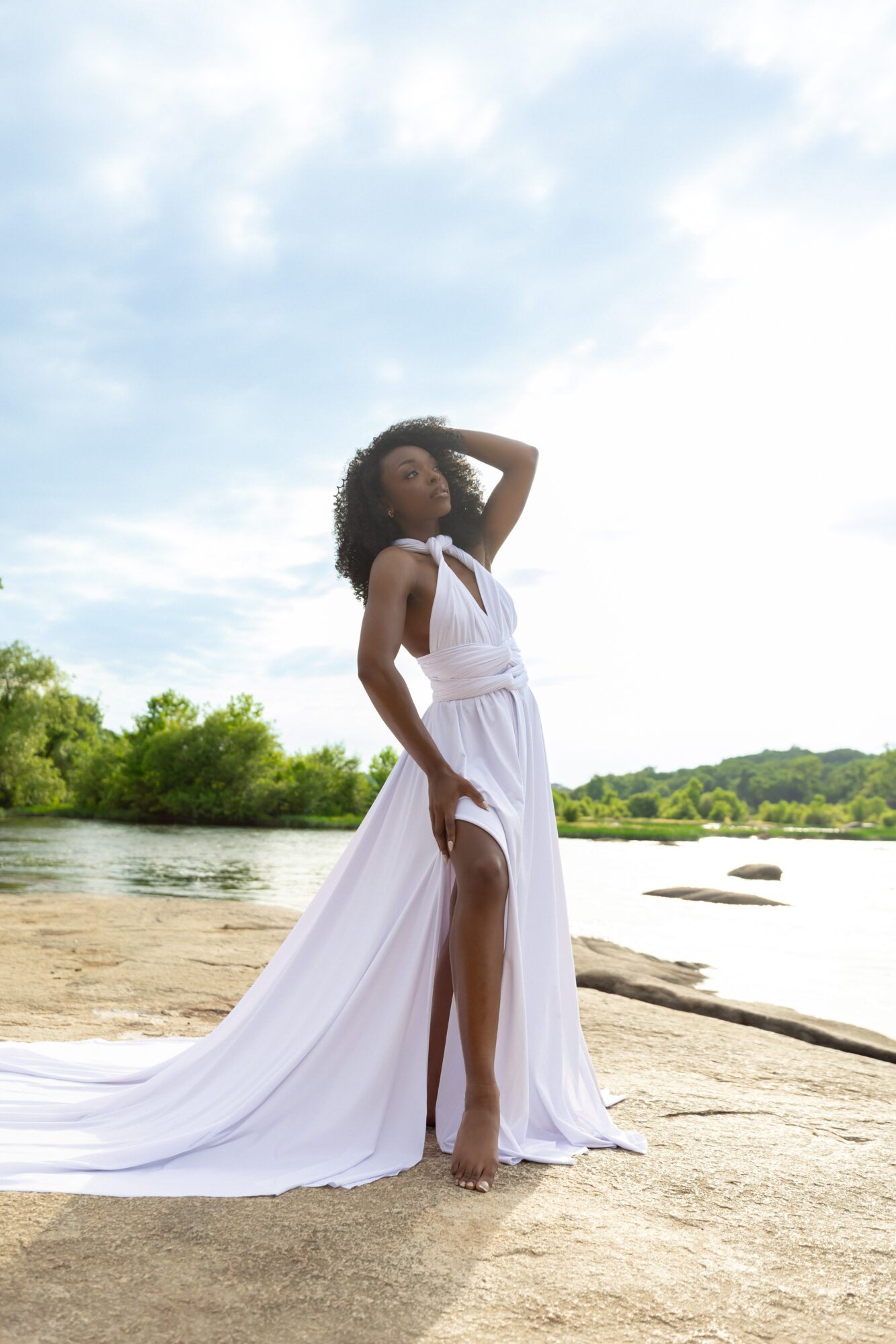 Woman in a white dress standing on a sandy riverbank with trees and water in the background, under a partly cloudy sky.